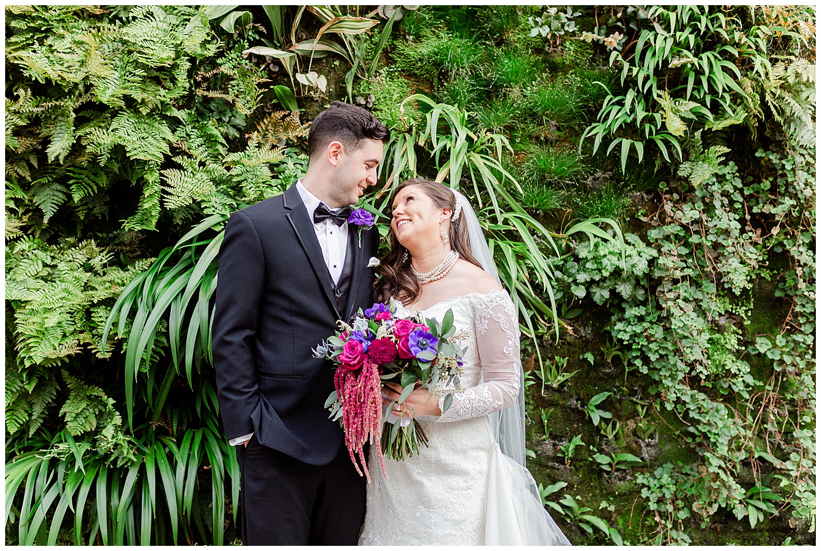 💍 bride groom outdoor wedding portraits in front of gorgeous wall of greenery florals and downtown city bridals 👰 Bride: lace wedding dress with rhinestone belt rhinestone barrette in hair down with pearls 🤵 Groom: black tux tuxedo and purple flower boutonnière 💍 Bright Colorful Summer City Wedding in Charlotte, NC with Taryn and Ryan | Kevyn Dixon Photography