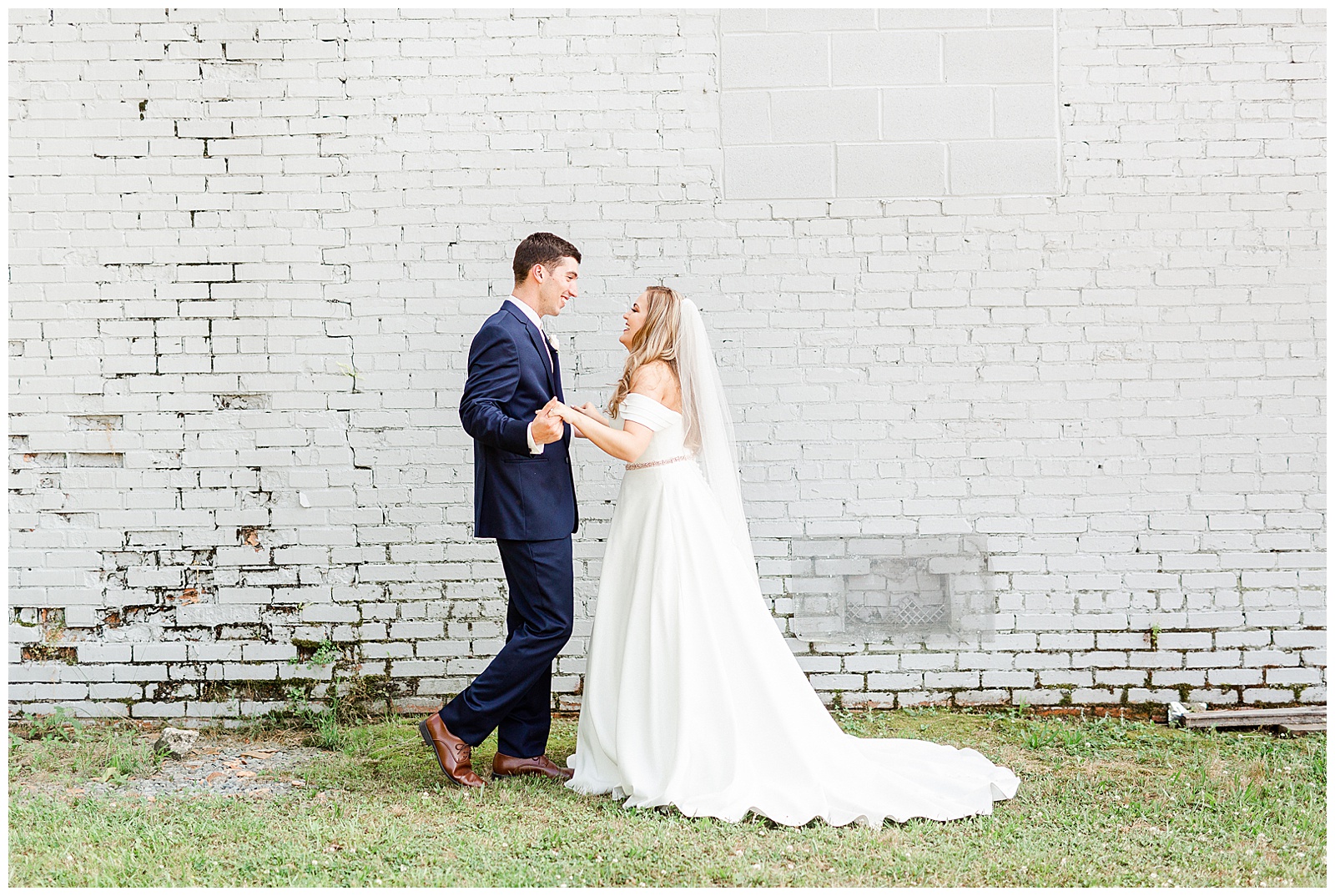 Stunning Modern Bride and Groom with off shoulder wedding dress and blue suit in outdoor portraits in front of white grungy wall from Summer Wedding in Charlotte, NC | Check out the full wedding at KevynDixonPhoto.com