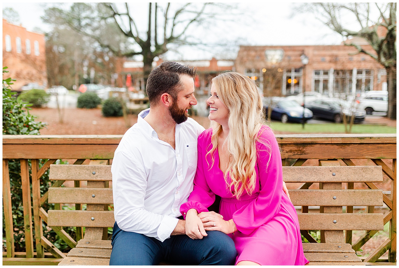 Adorable couple sitting on wooden bench in downtown Waxhaw, NC Engagement Session with Bright Hot Pink Dress Outfit Ideas