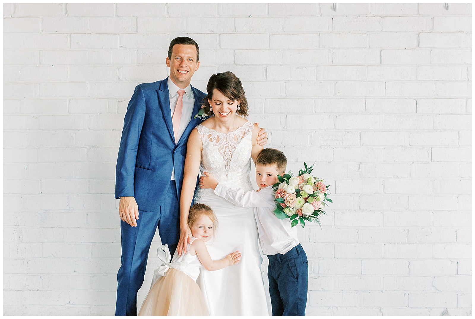 adorable shot with flower girl and ring bearer from indoor summer bride groom portraits of short brown haired bride in lace dress with long train and navy blue suited groom