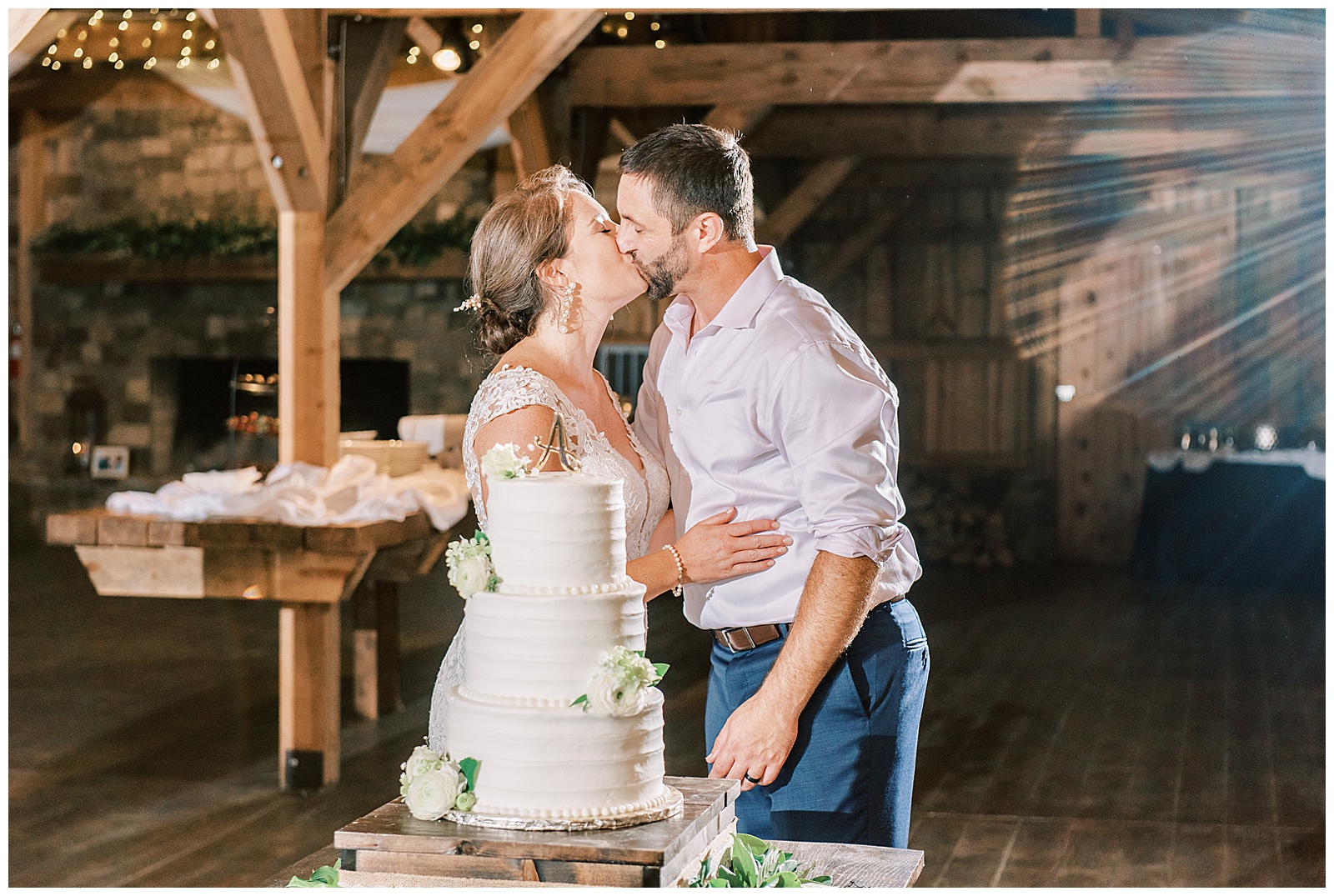 bride and groom kiss in front of three tiered white cake at indoor summer wedding reception in wooden barn