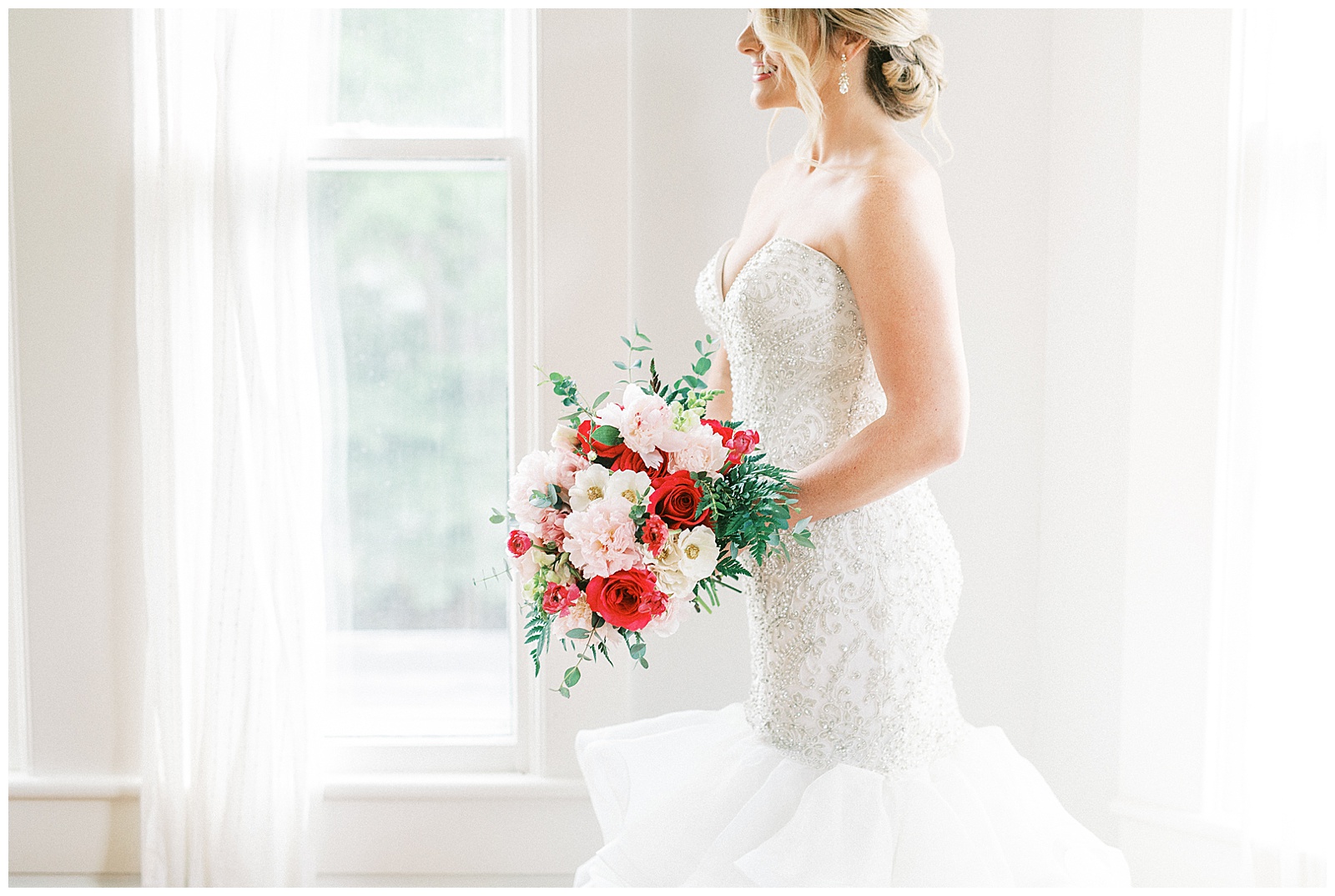 gorgeous light and airy indoor bridal portraits of blonde haired bride in mermaid dress with loads of tulle next to floor length window with red and white bouquet