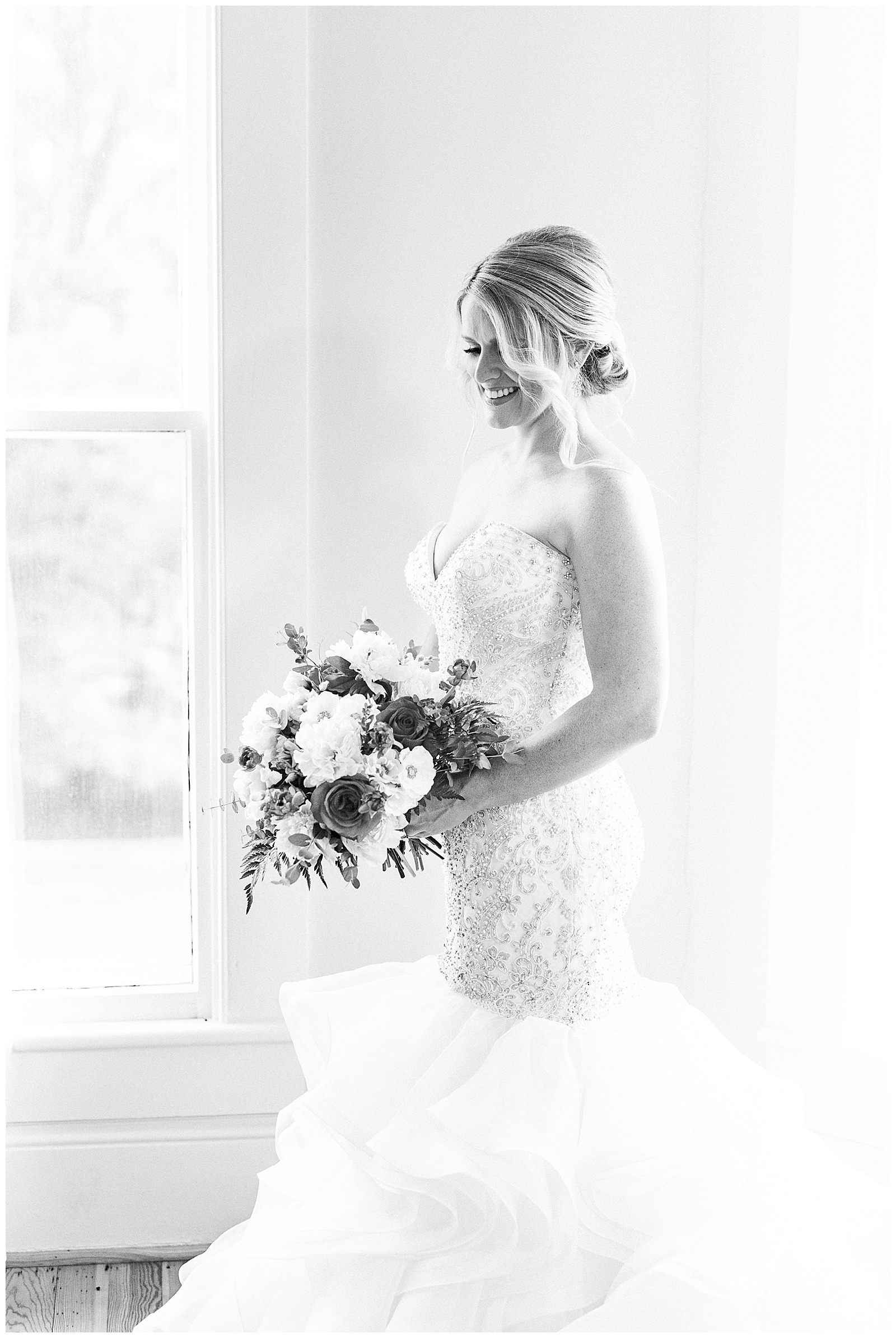 gorgeous light and airy indoor bridal portraits of blonde haired bride in mermaid dress with loads of tulle next to floor length window with red and white bouquet