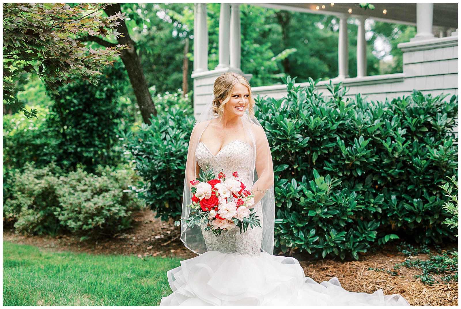 gorgeous light and airy outdoor bridal portrait on southern front porch of blonde haired bride in mermaid dress with loads of tulle with red and white bouquet