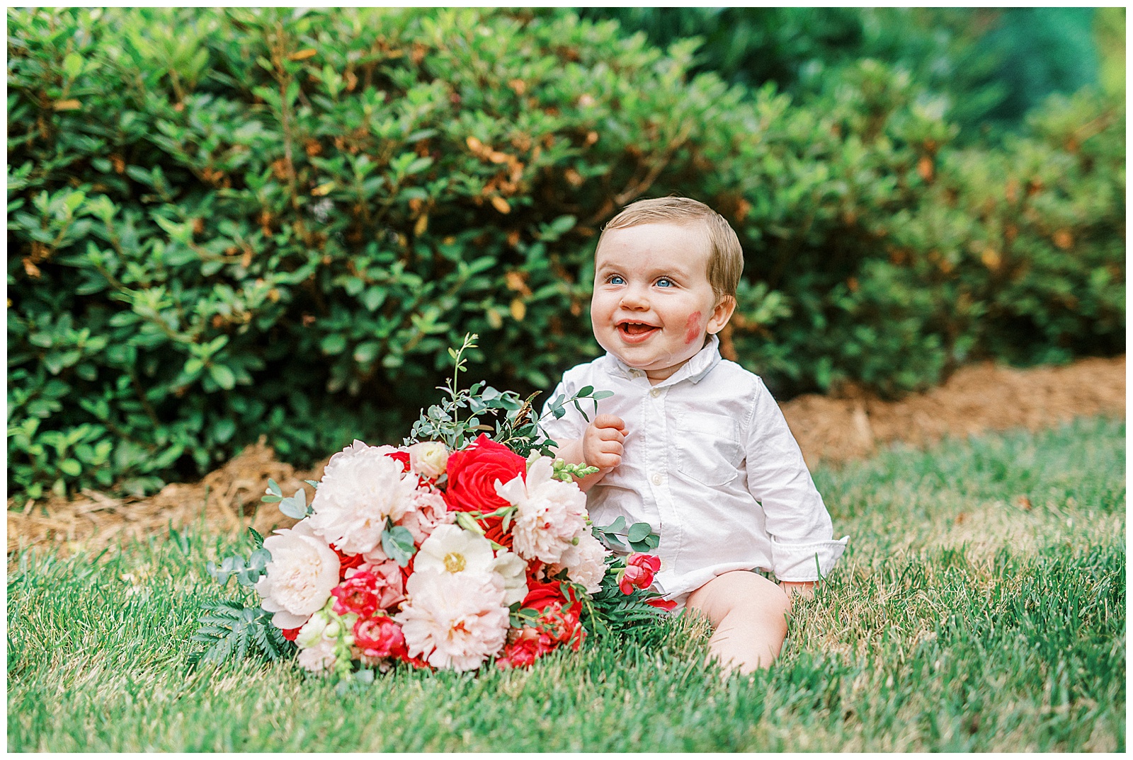 adorable baby son of bride poses with red and white bouquet and lipstick kiss on cheek outdoor portrait in the grass