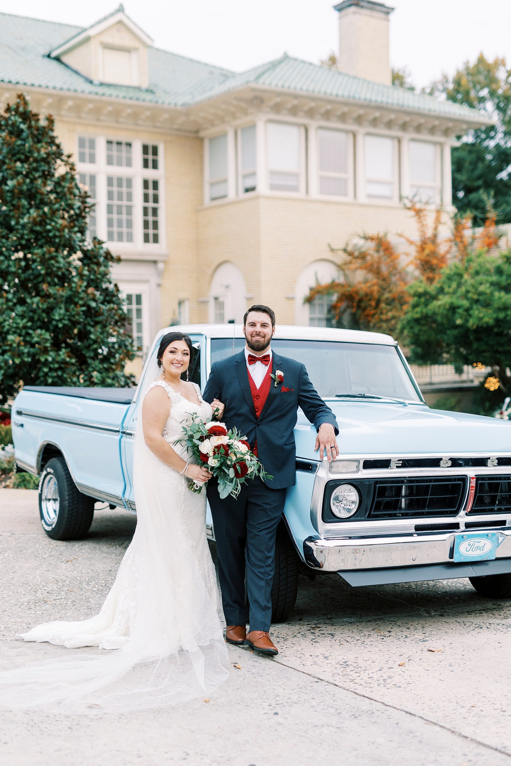 groom leans against hood of resorted blue Ford truck with bride standing next to him