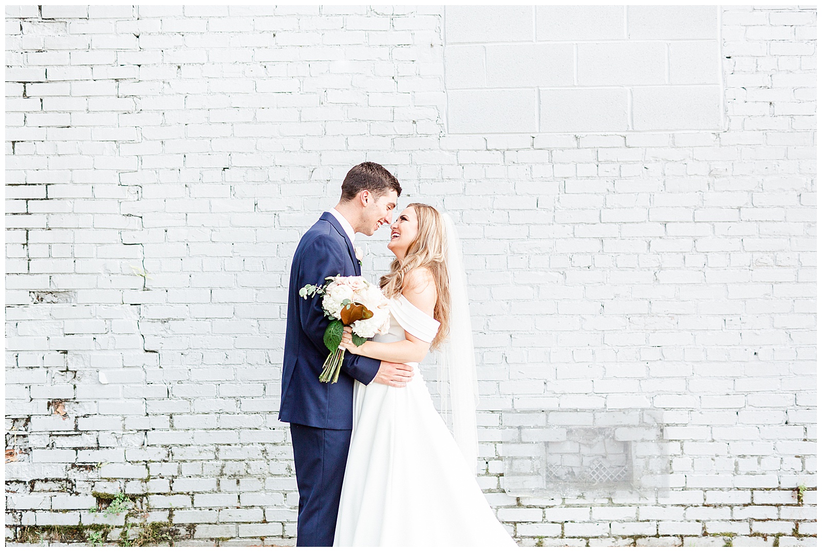 Stunning Modern Bride and Groom with off shoulder wedding dress and blue suit in outdoor portraits in front of white grungy wall from Summer Wedding in Charlotte, NC | Check out the full wedding at KevynDixonPhoto.com