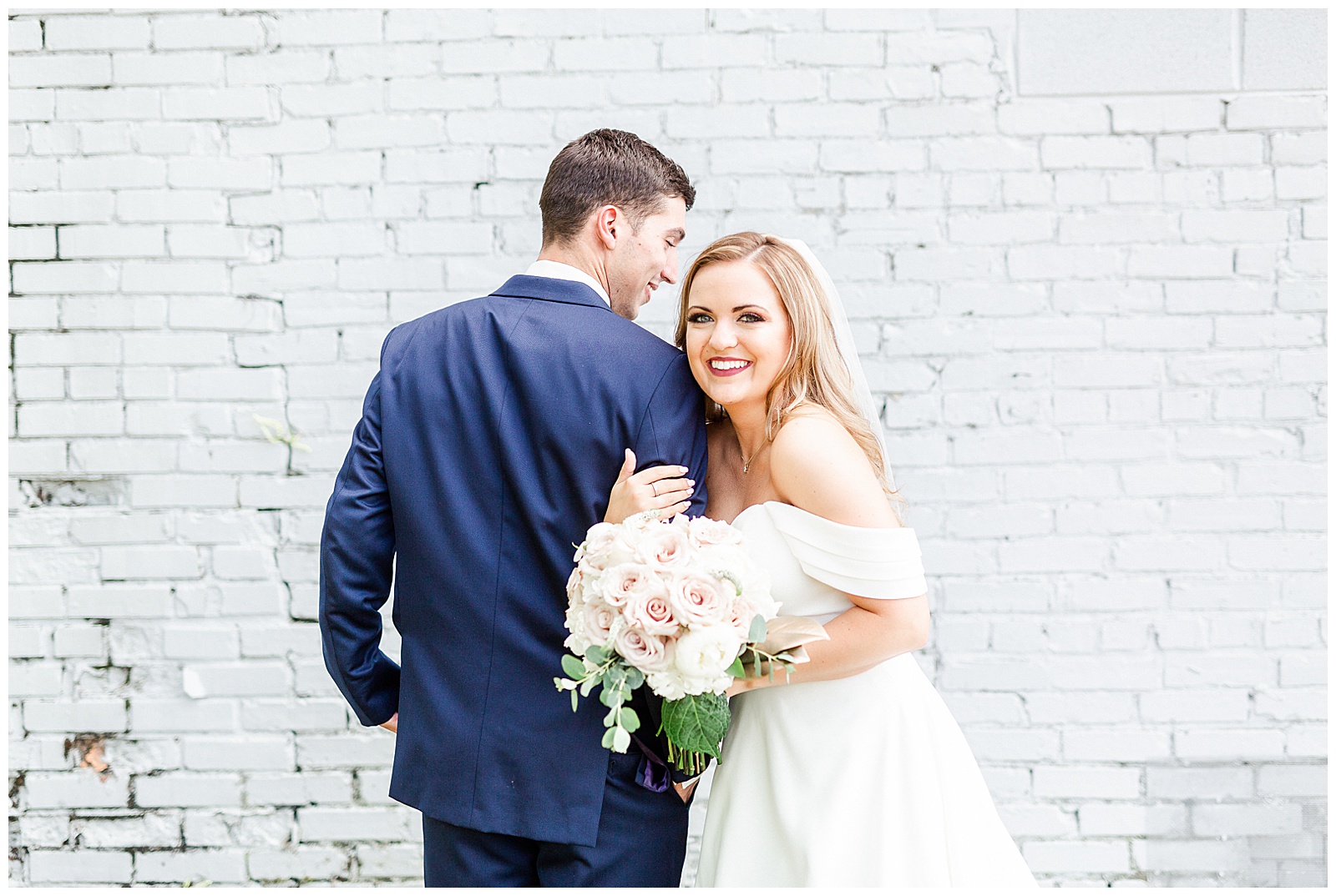 Stunning Modern Bride and Groom with off shoulder wedding dress and blue suit in outdoor portraits in front of white grungy wall from Summer Wedding in Charlotte, NC | Check out the full wedding at KevynDixonPhoto.com