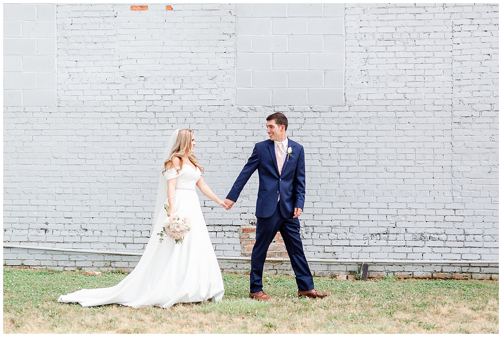 Stunning Modern Bride and Groom with off shoulder wedding dress and blue suit in outdoor portraits in front of white grungy wall from Summer Wedding in Charlotte, NC | Check out the full wedding at KevynDixonPhoto.com