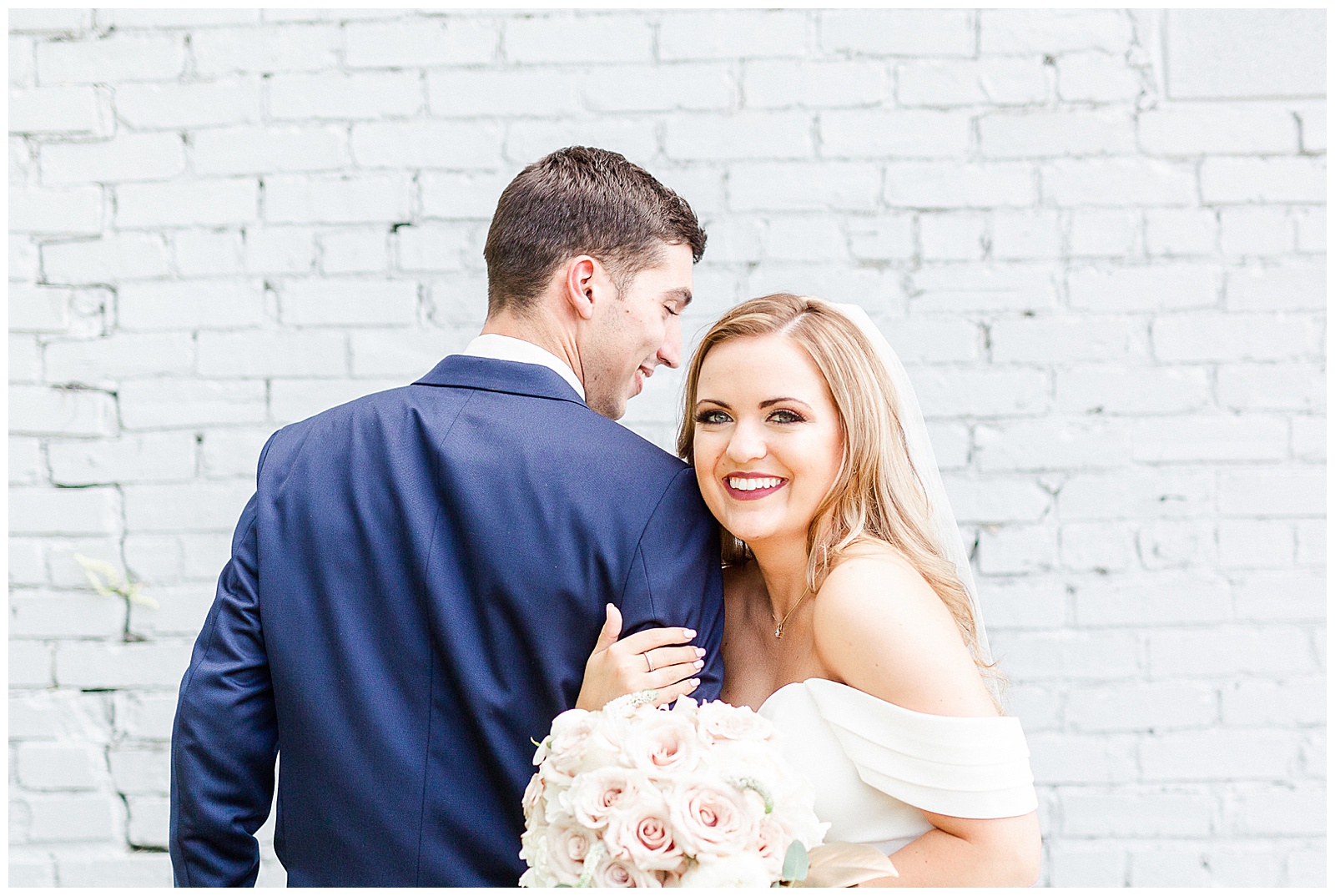 Stunning Modern Bride and Groom with off shoulder wedding dress and blue suit in outdoor portraits in front of white grungy wall from Summer Wedding in Charlotte, NC | Check out the full wedding at KevynDixonPhoto.com