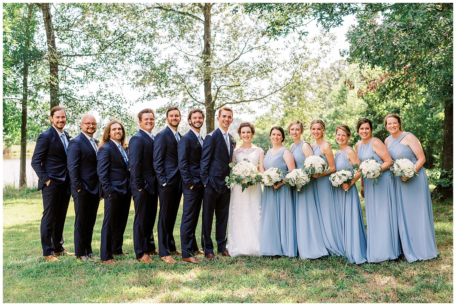 dusty blue bridesmaid bride tribe with white flower bouquet and navy blue suit groomsmen summer outdoor bridal party group portrait