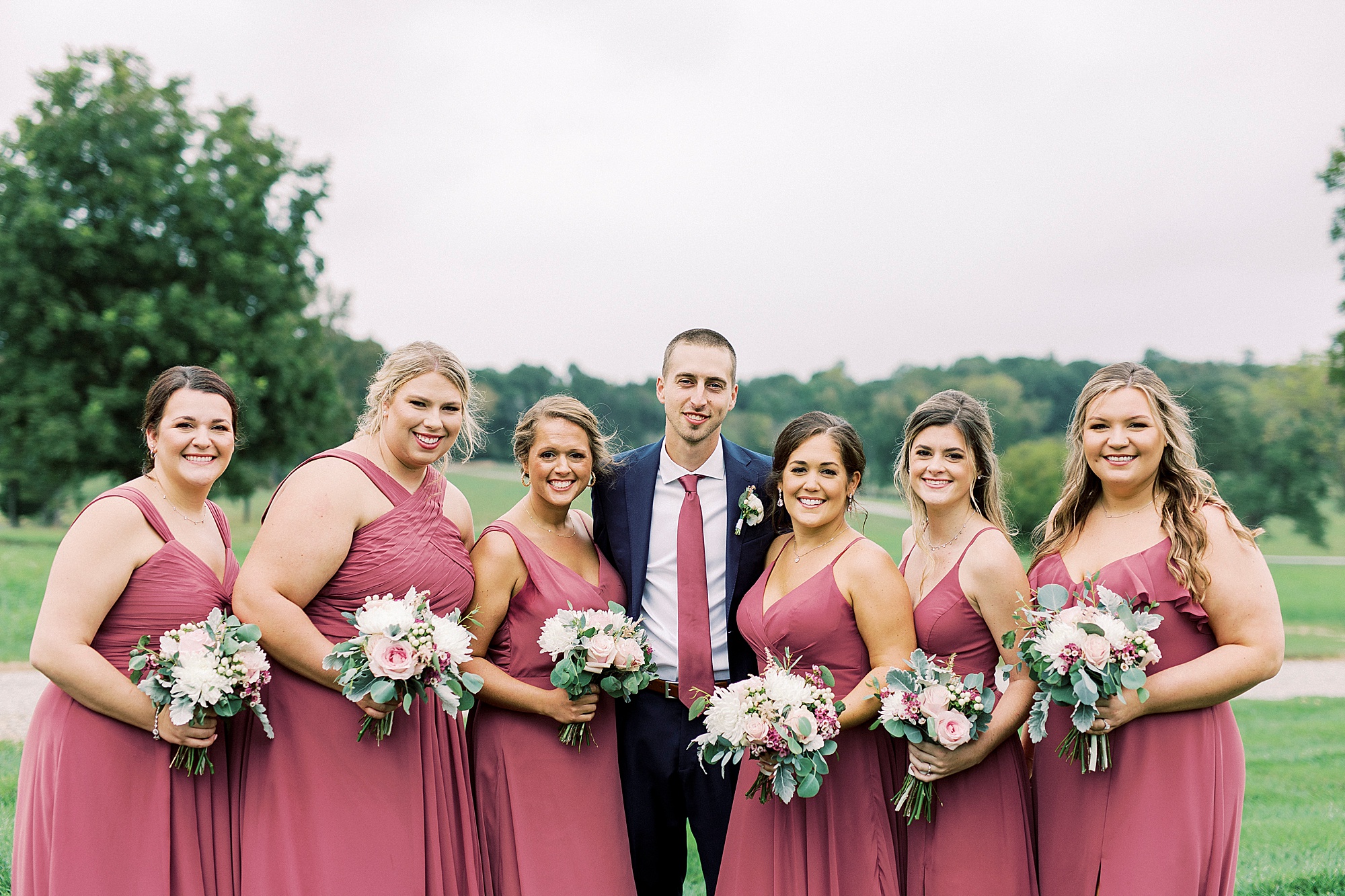 groom poses with bridesmaids in dark pink gowns