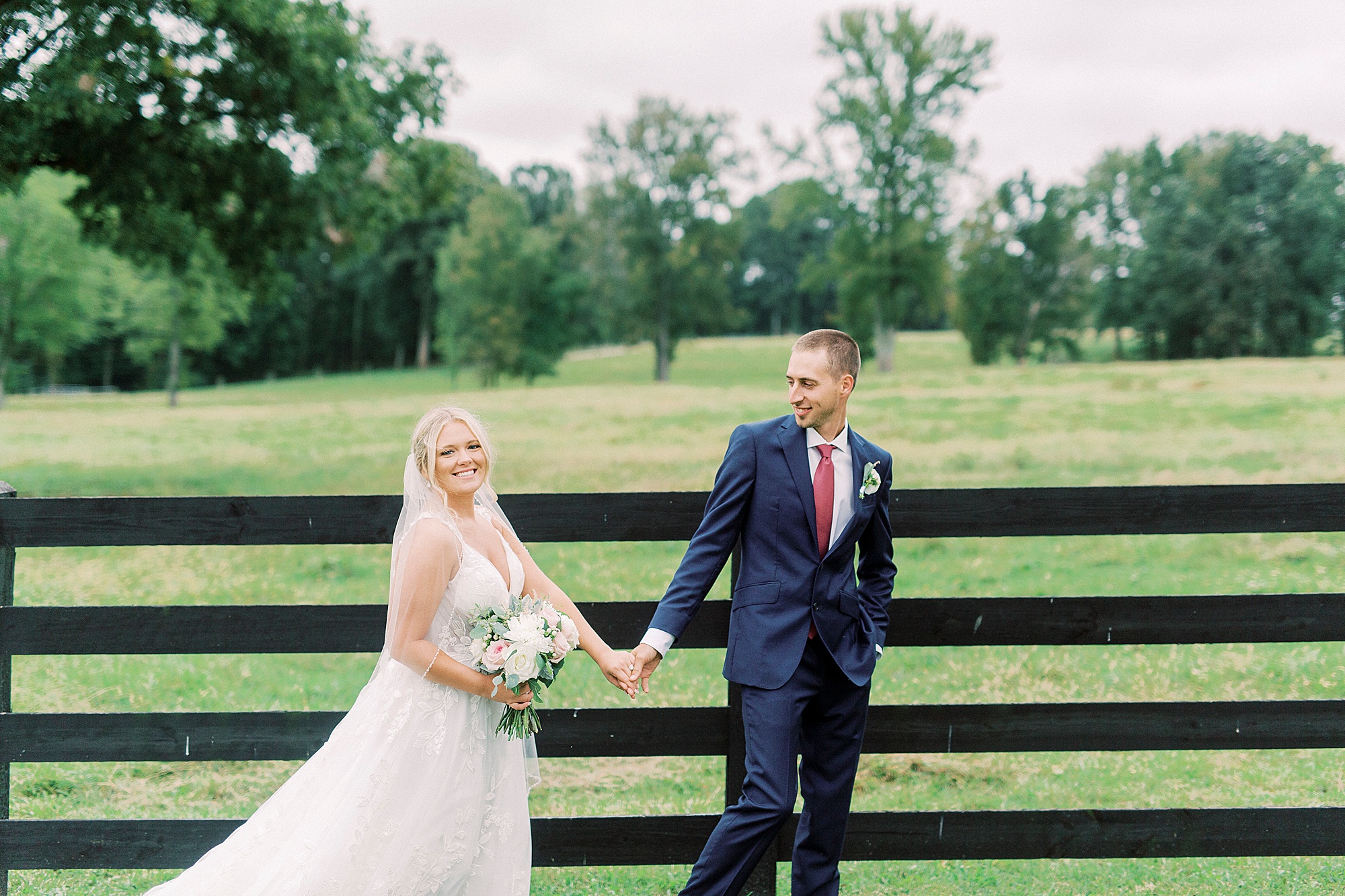 newlyweds walk by dark wooden fence at Addison Farm