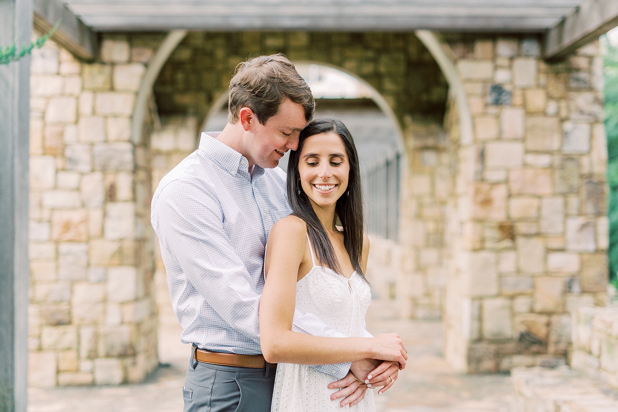engaged couple stands under stone archway in Midtown Park
