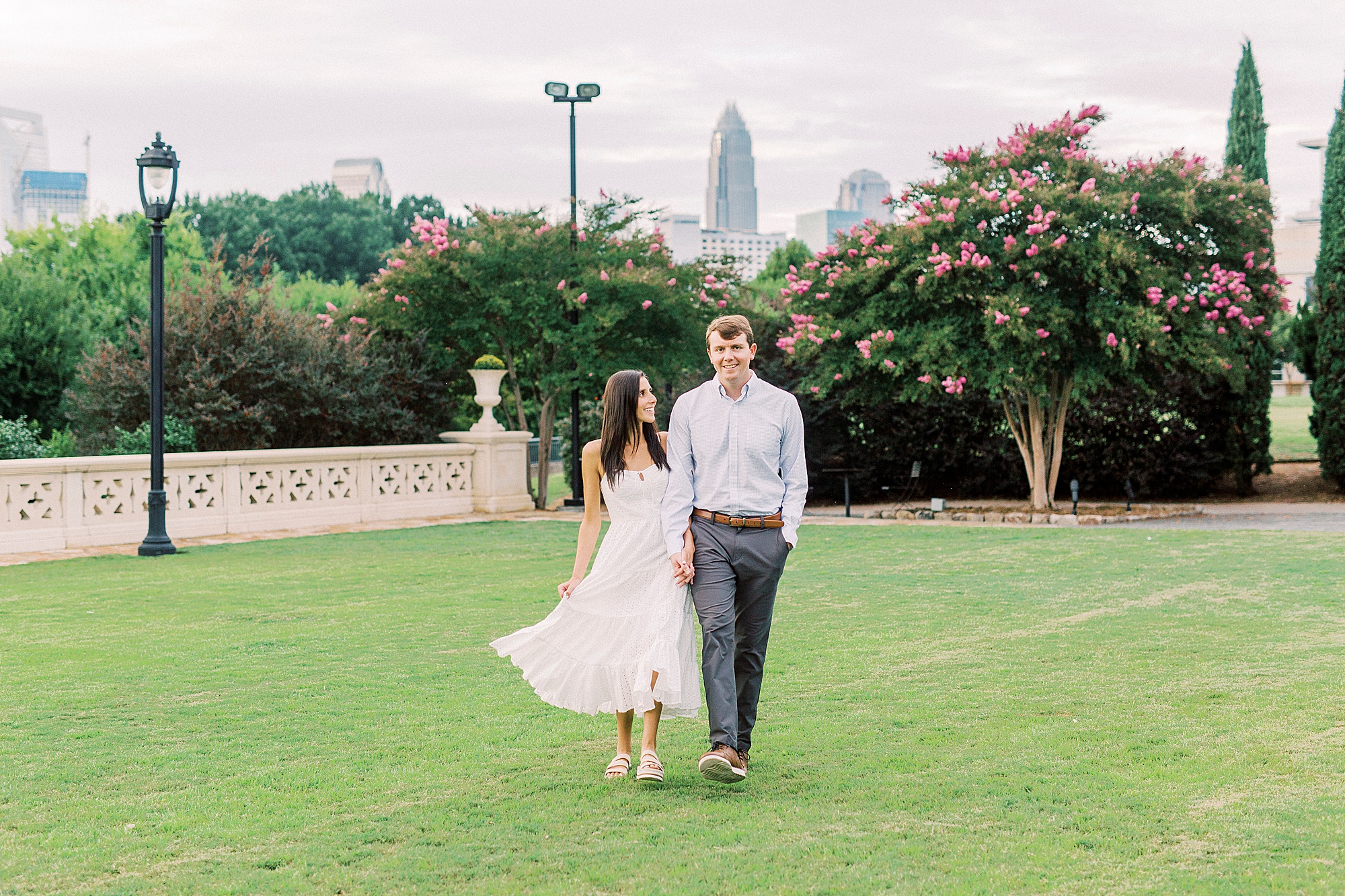 couple walks through Midtown Park with Charlotte skyline behind them