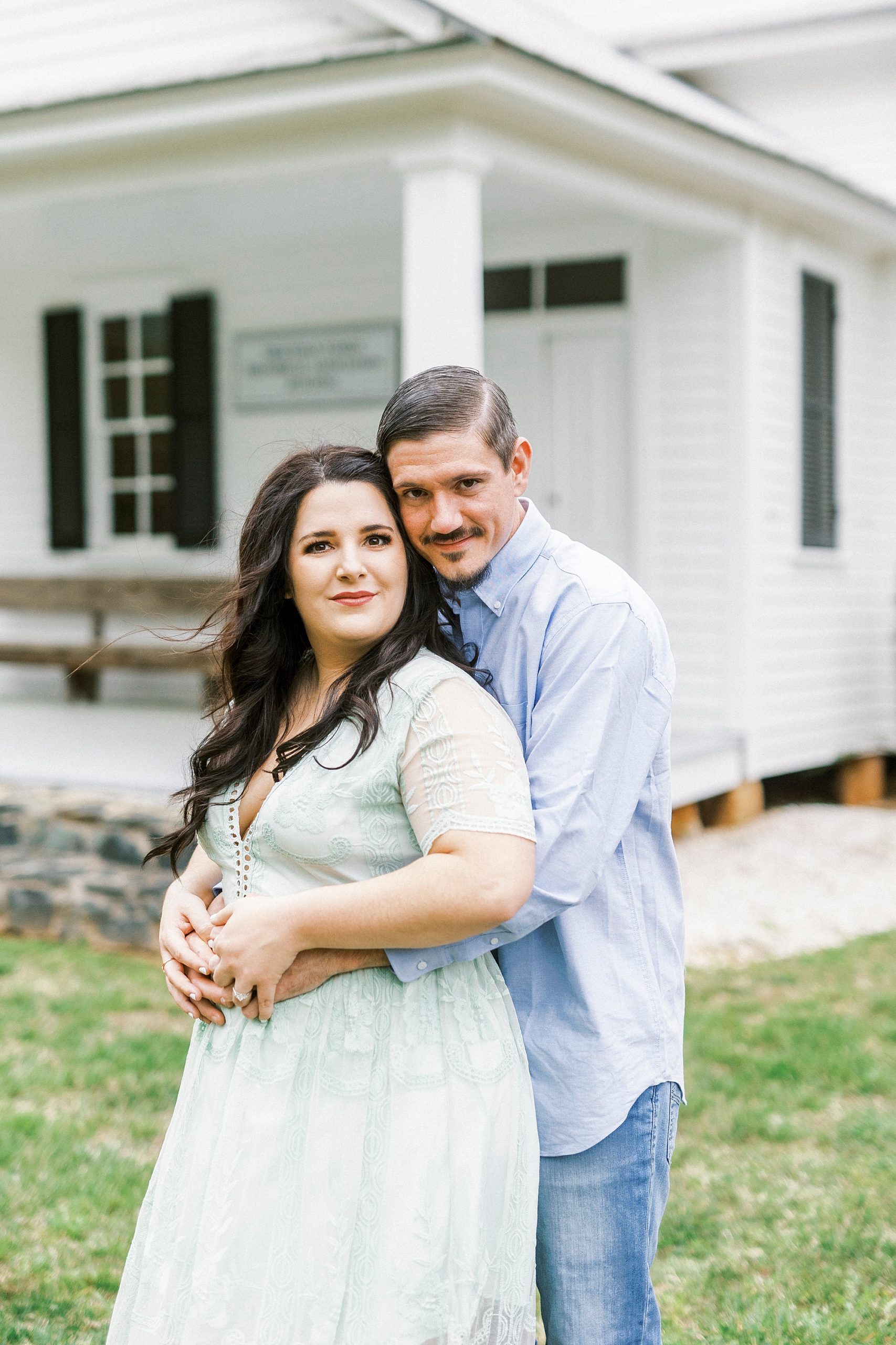 engaged couple hugs in yard of white chapel 