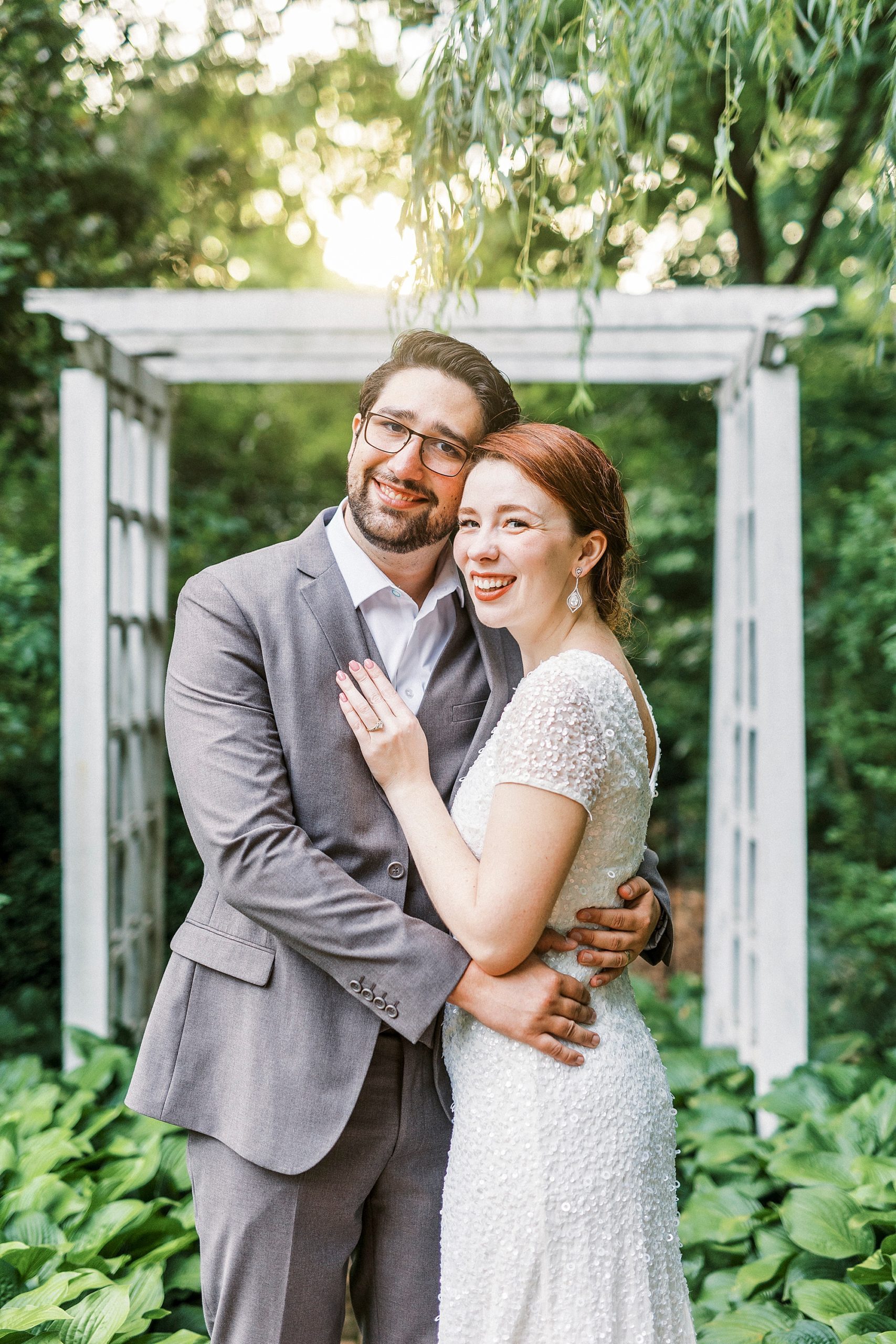 couple hugs under white arbor at Wing Haven Garden