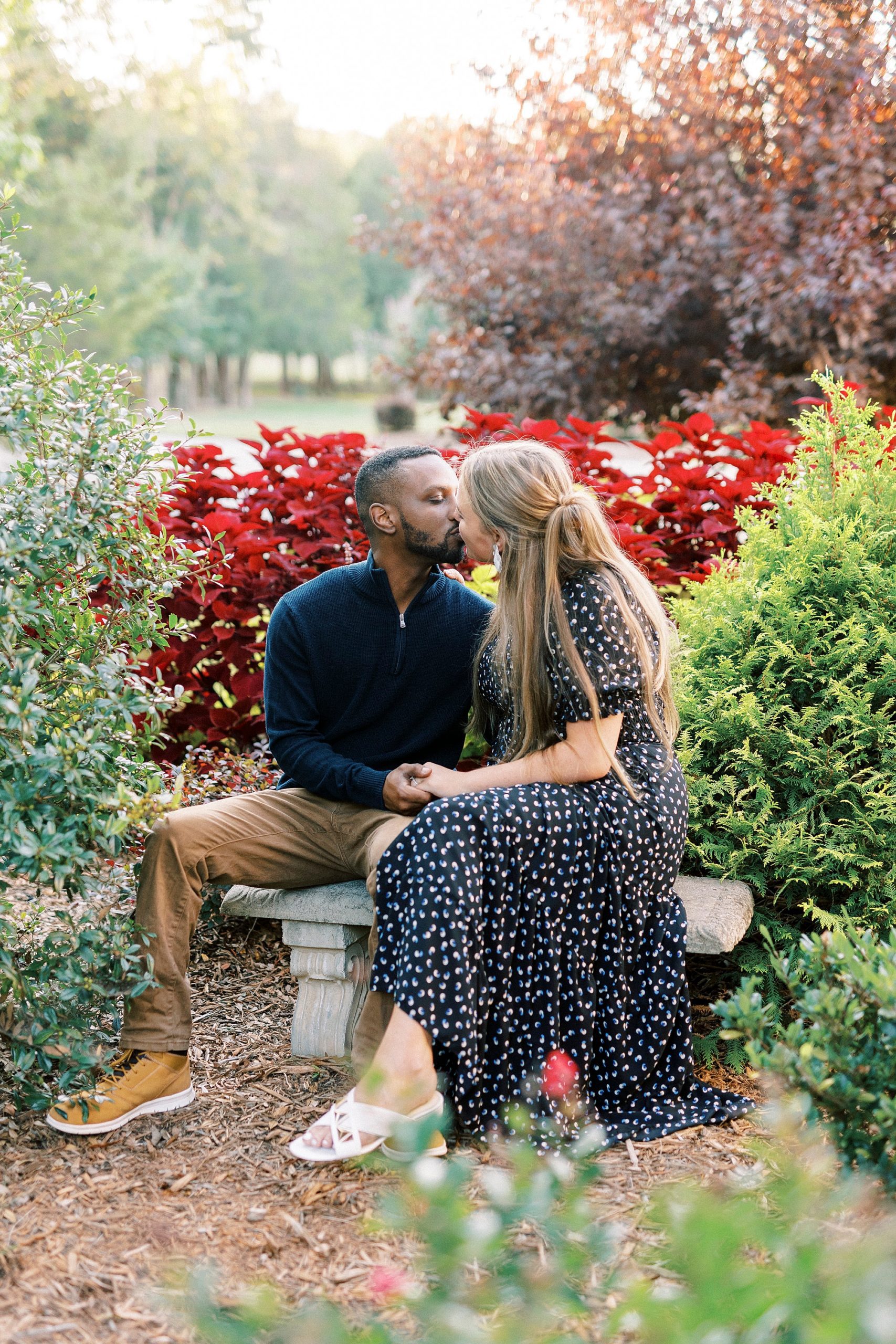 engaged couple kisses on stone bench in gardens at Bella Terra