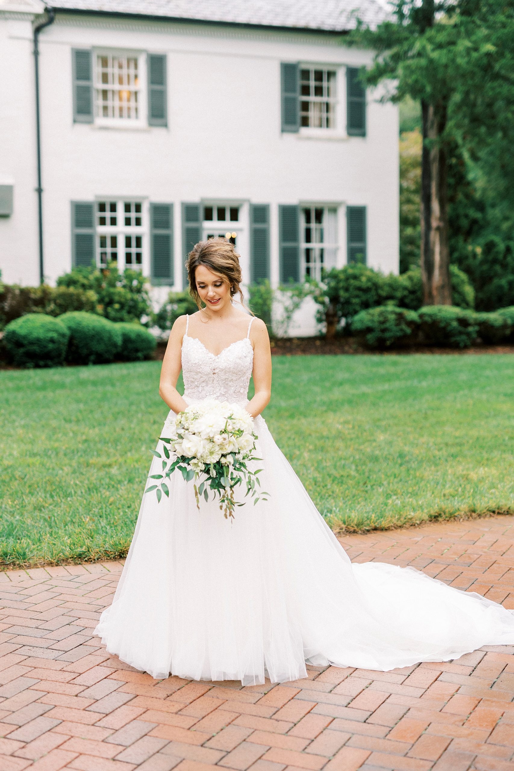 bride stands in wedding gown with spaghetti straps looking down at bouquet 