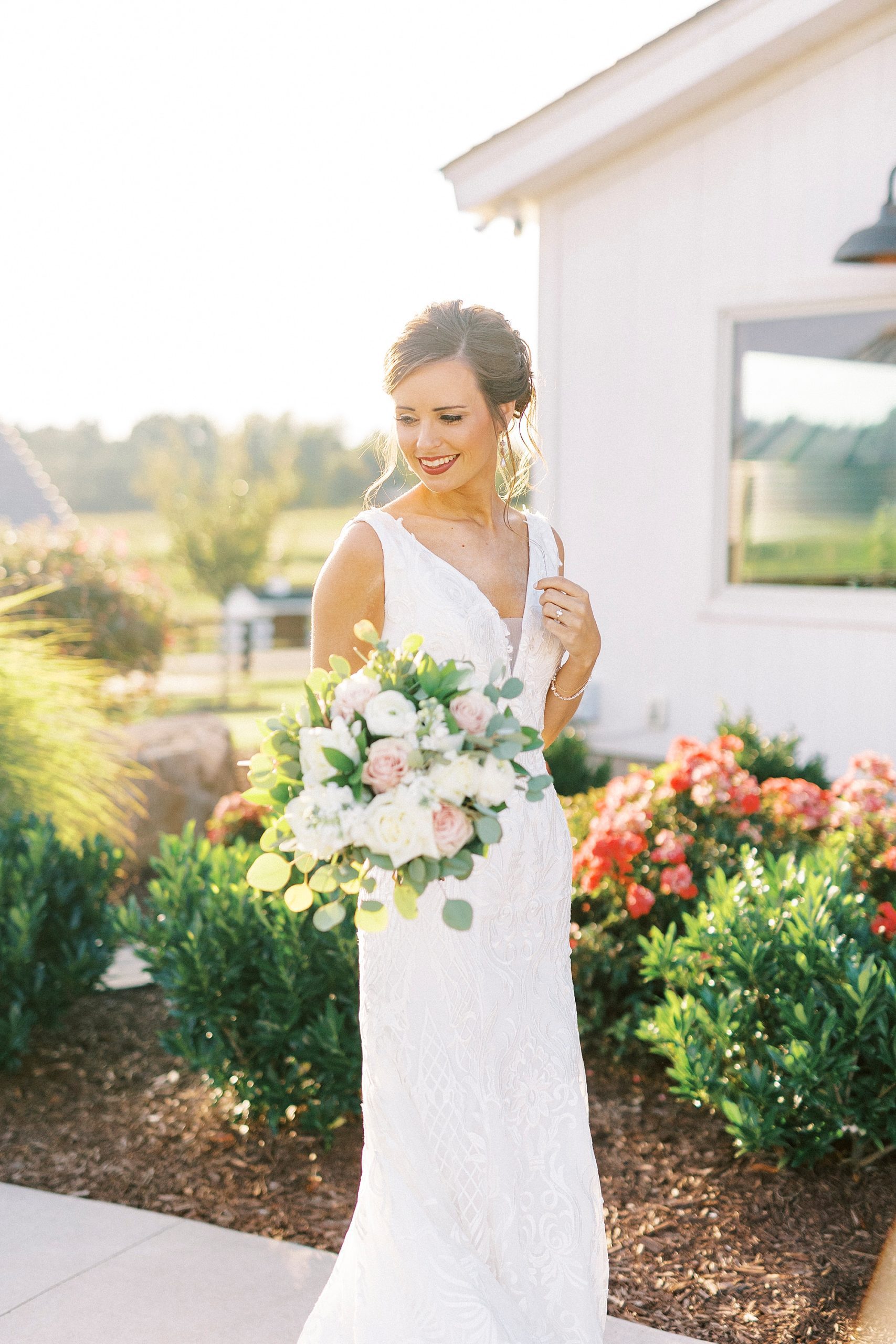 bride poses by white barn looking over bouquet
