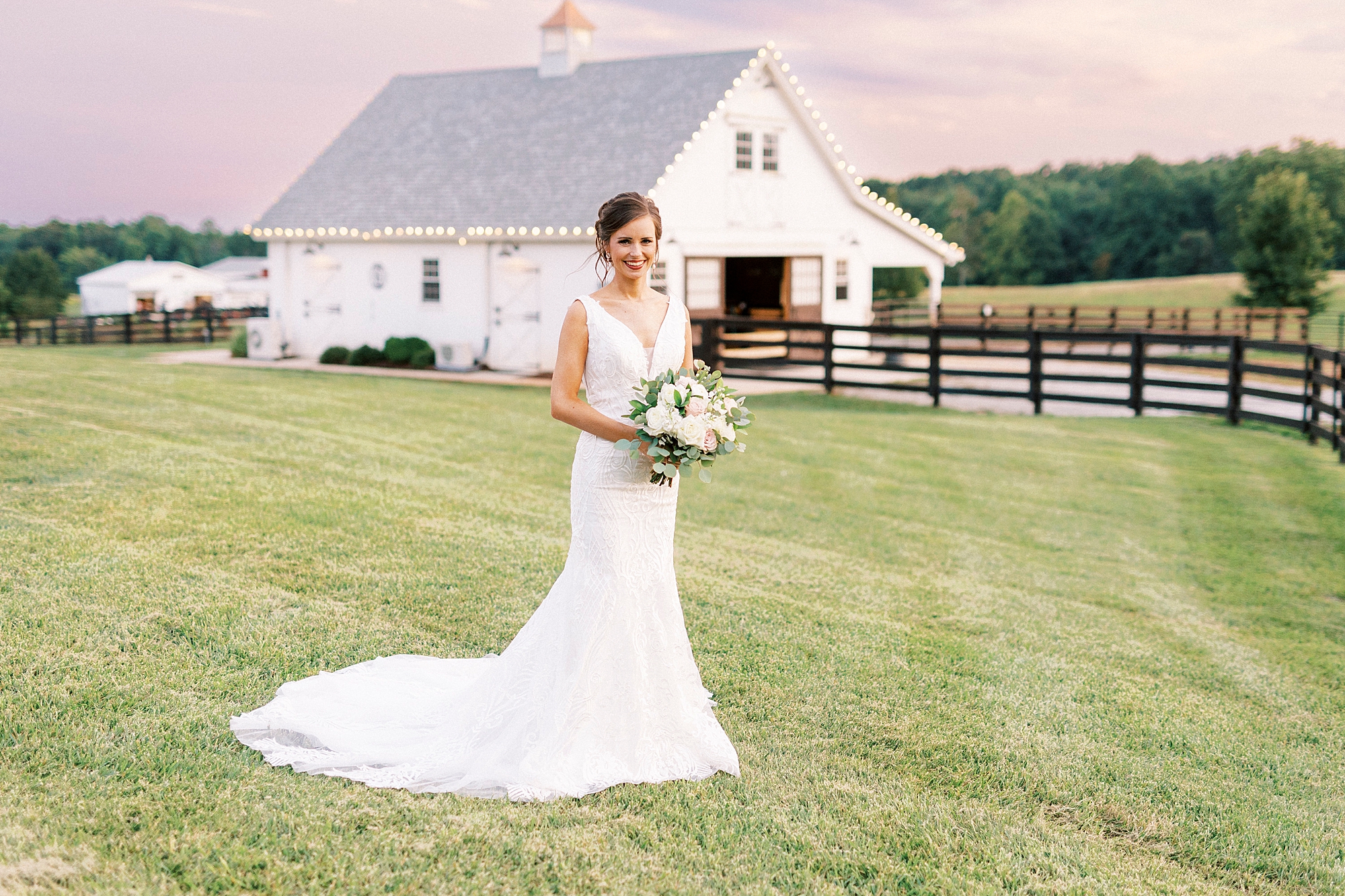 bride stands in front of white barn at sunset