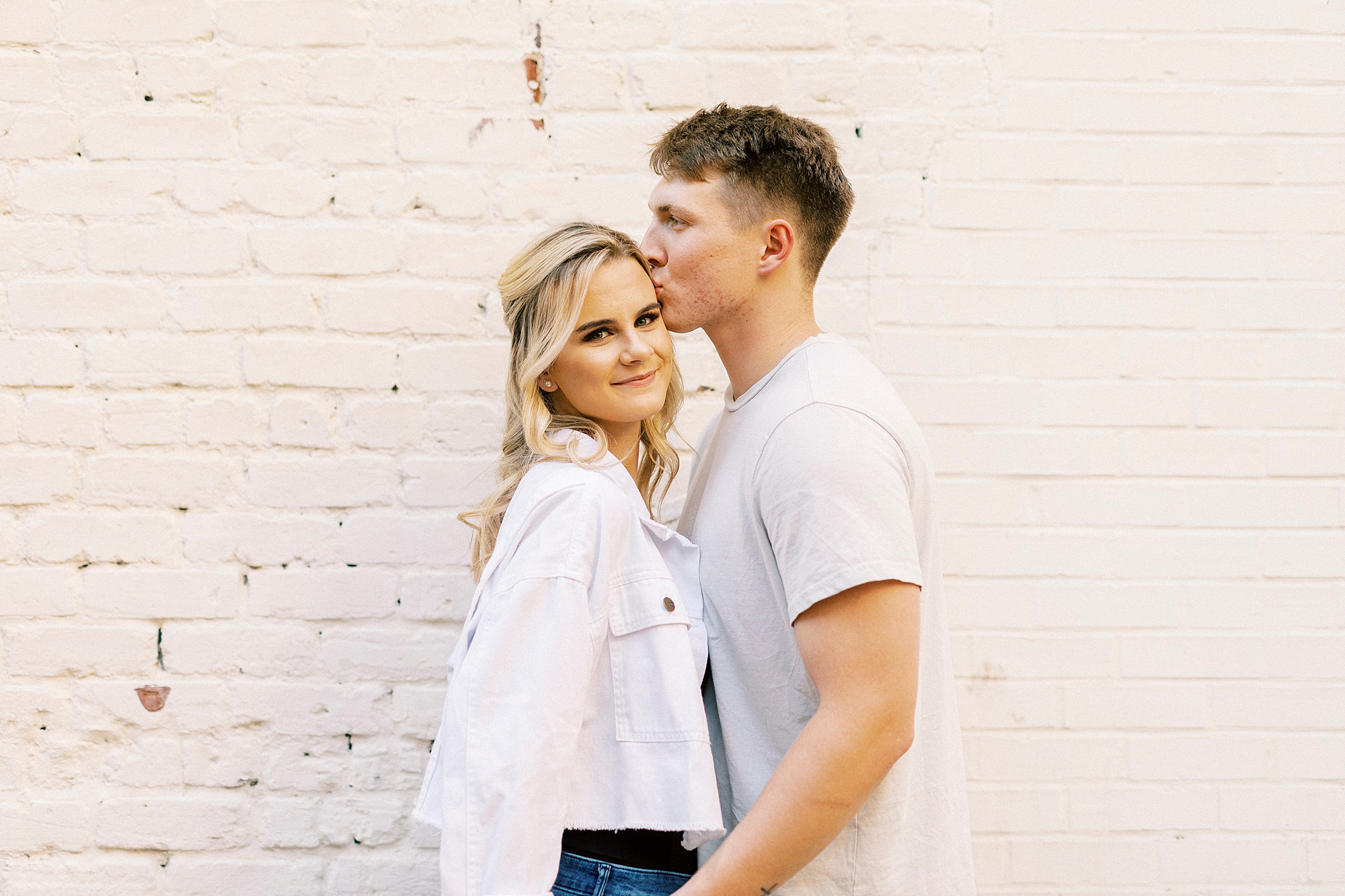 groom kisses fiancee's head in front of white wall during Downtown Concord engagement portraits