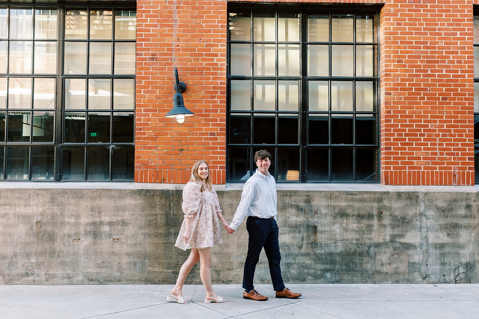 engaged couple holds hands walking along brick warehouse at the American Tobacco Campus
