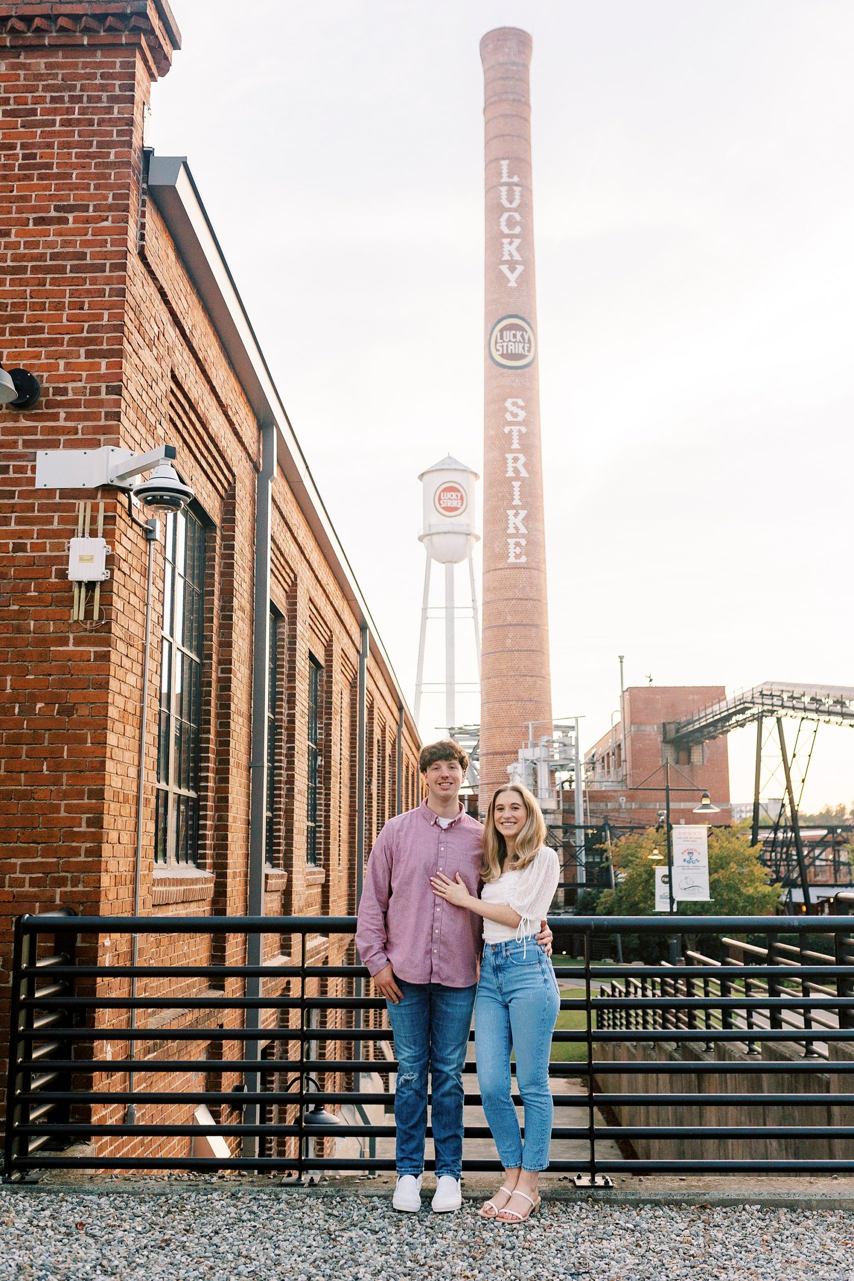 couple hugs with smoke stack at American Tobacco Campus behind them