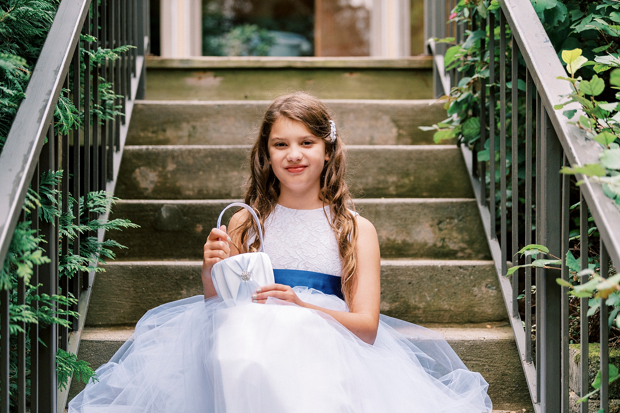 flower girl sits on step in white dress with blue satin ribbon