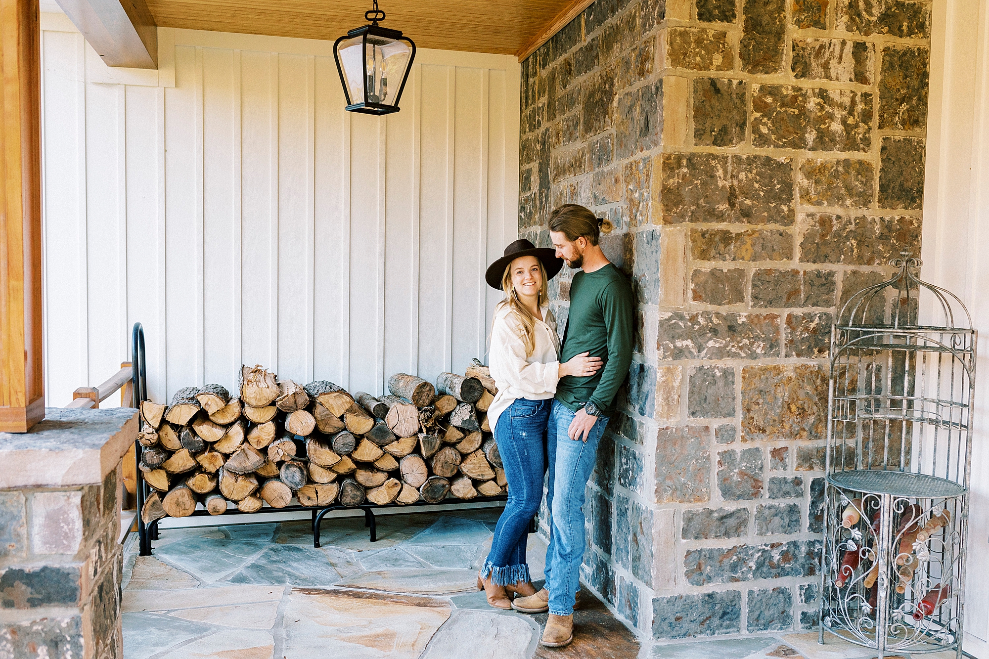 engaged couple hugs against stone wall near woodpile at home