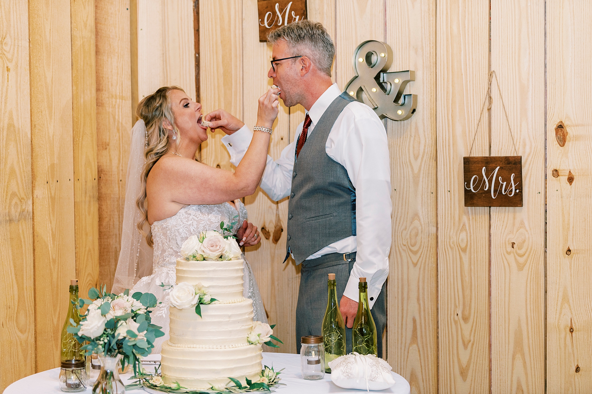bride and groom feed each other cake by wooden wall of barn