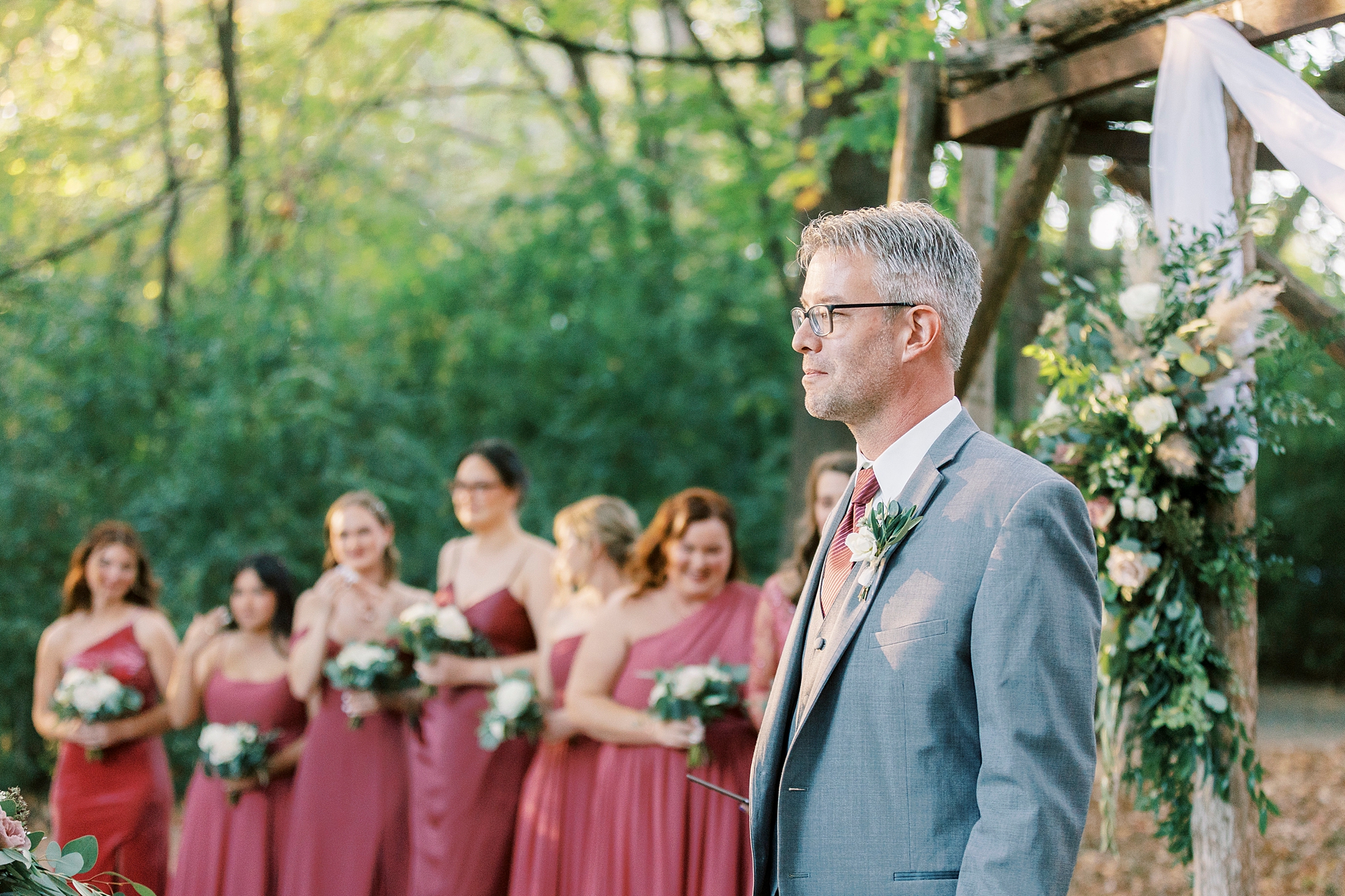 groom stands in grey suit by wooden arbor watching bride enter wedding ceremony