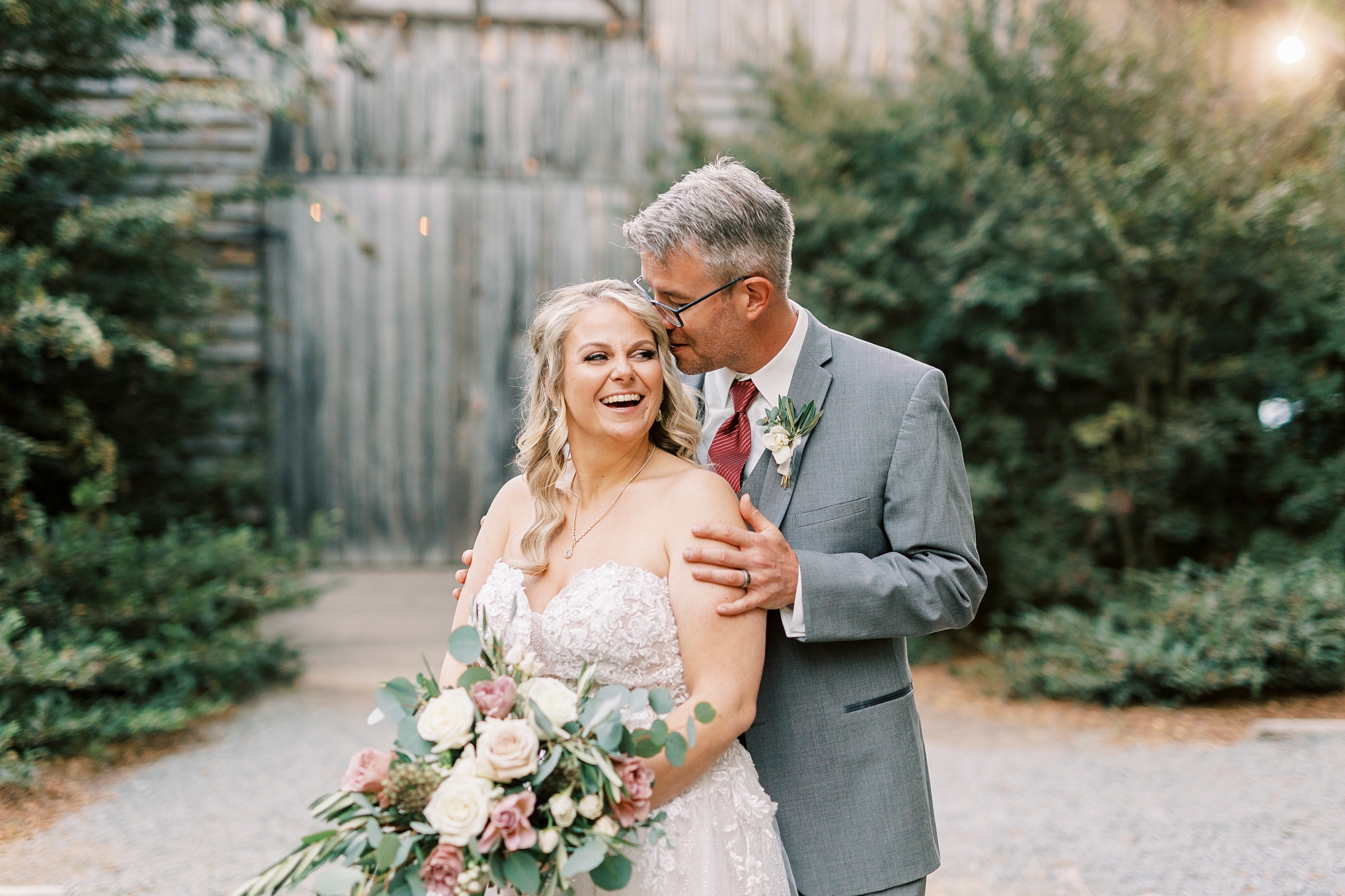 groom stands behind bride in grey suit and kisses her temple