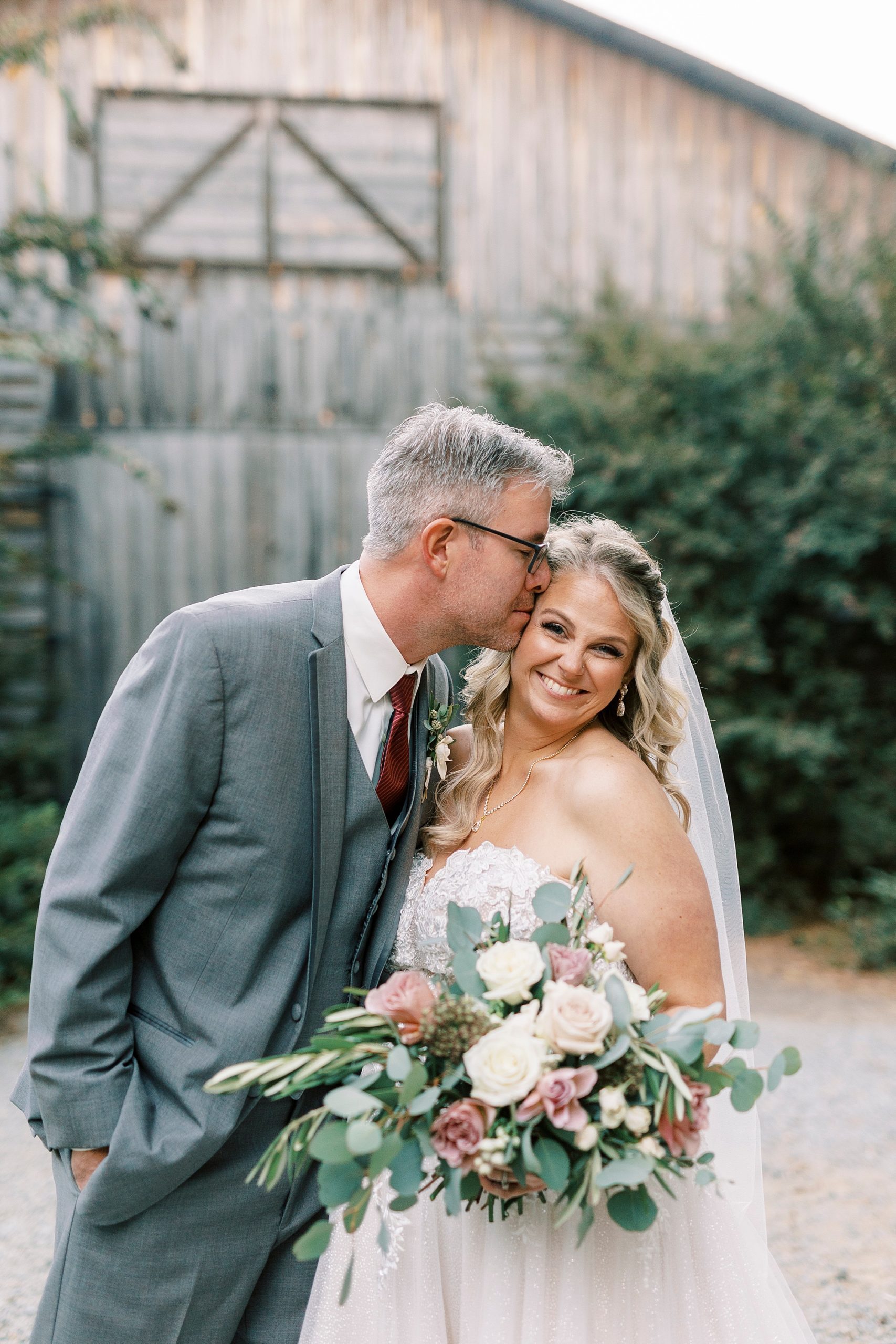 groom leans into bride kissing her forehead during portraits outside barn at the Gin at Aw Shucks Farm