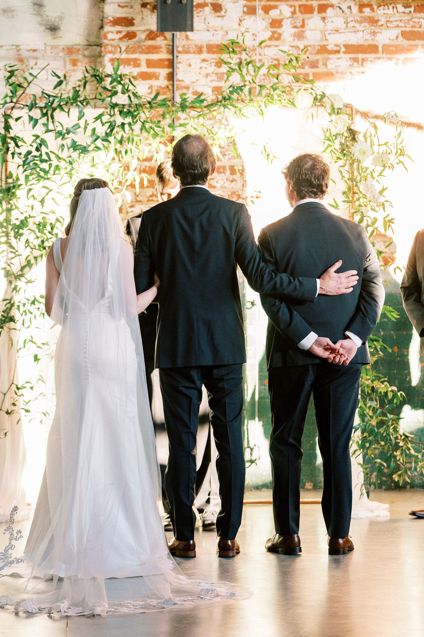 father of bride holds bride and hugs groom at alter before the Cadillac Service Garage for Christ-centered wedding ceremony in Greensboro NC 