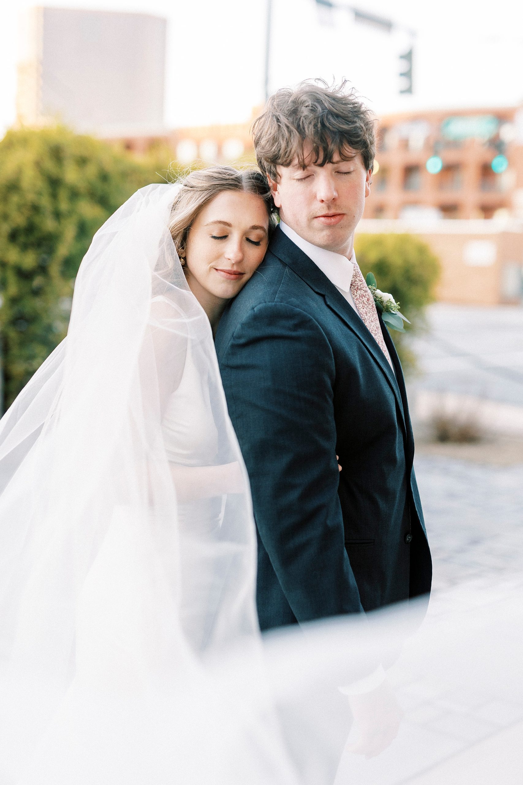 bride stands behind groom in navy suit, hugging him and laying head on his shoulders 