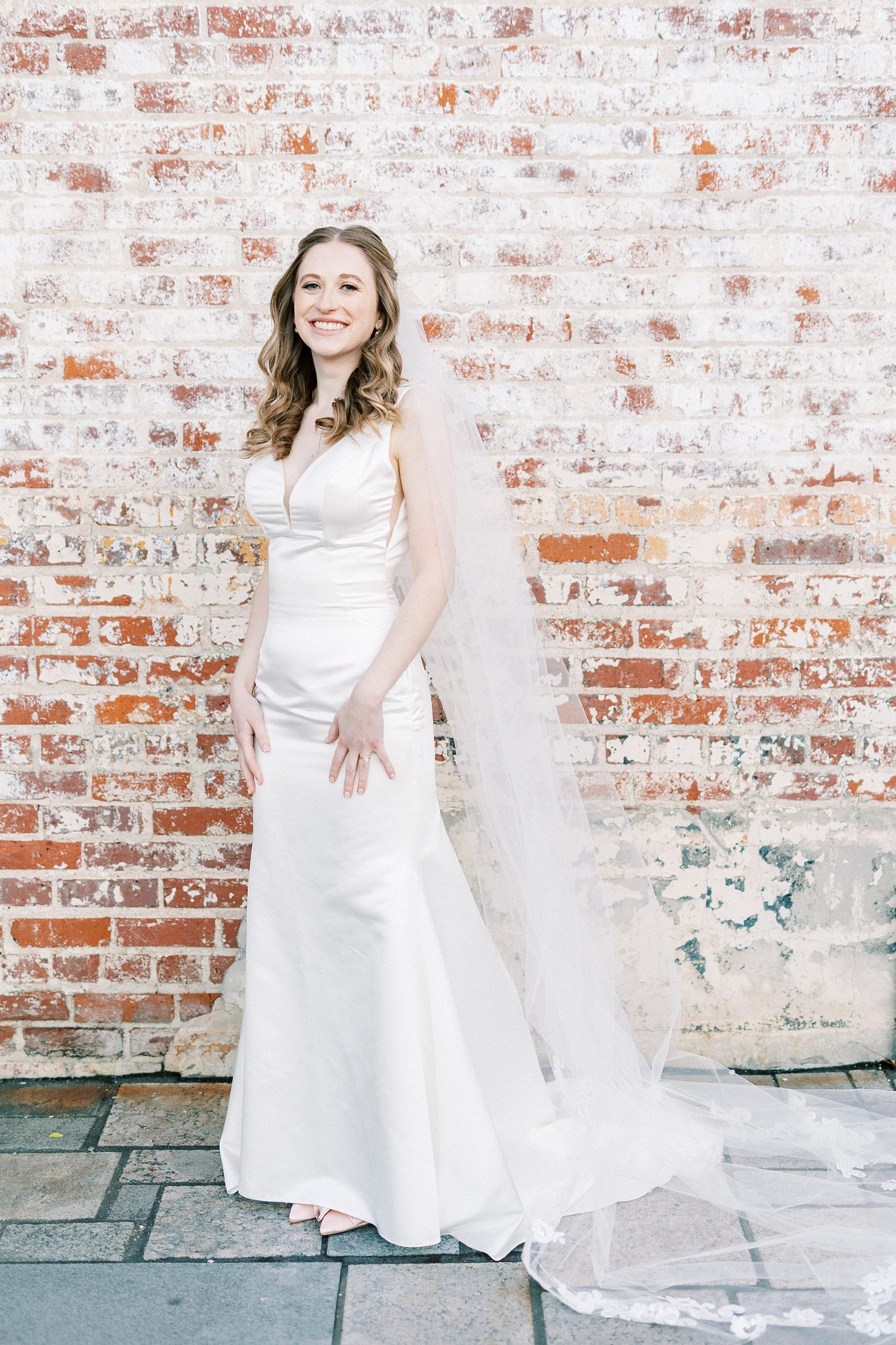 bride poses in wedding gown with veil around shoulders by brick wall at the Cadillac Service Garage