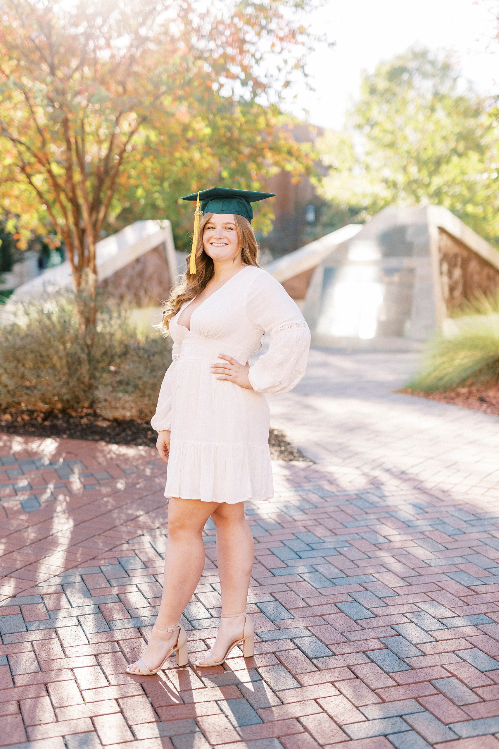 senior stands on brick patio in white dress and green cap during UNC Charlotte senior portraits