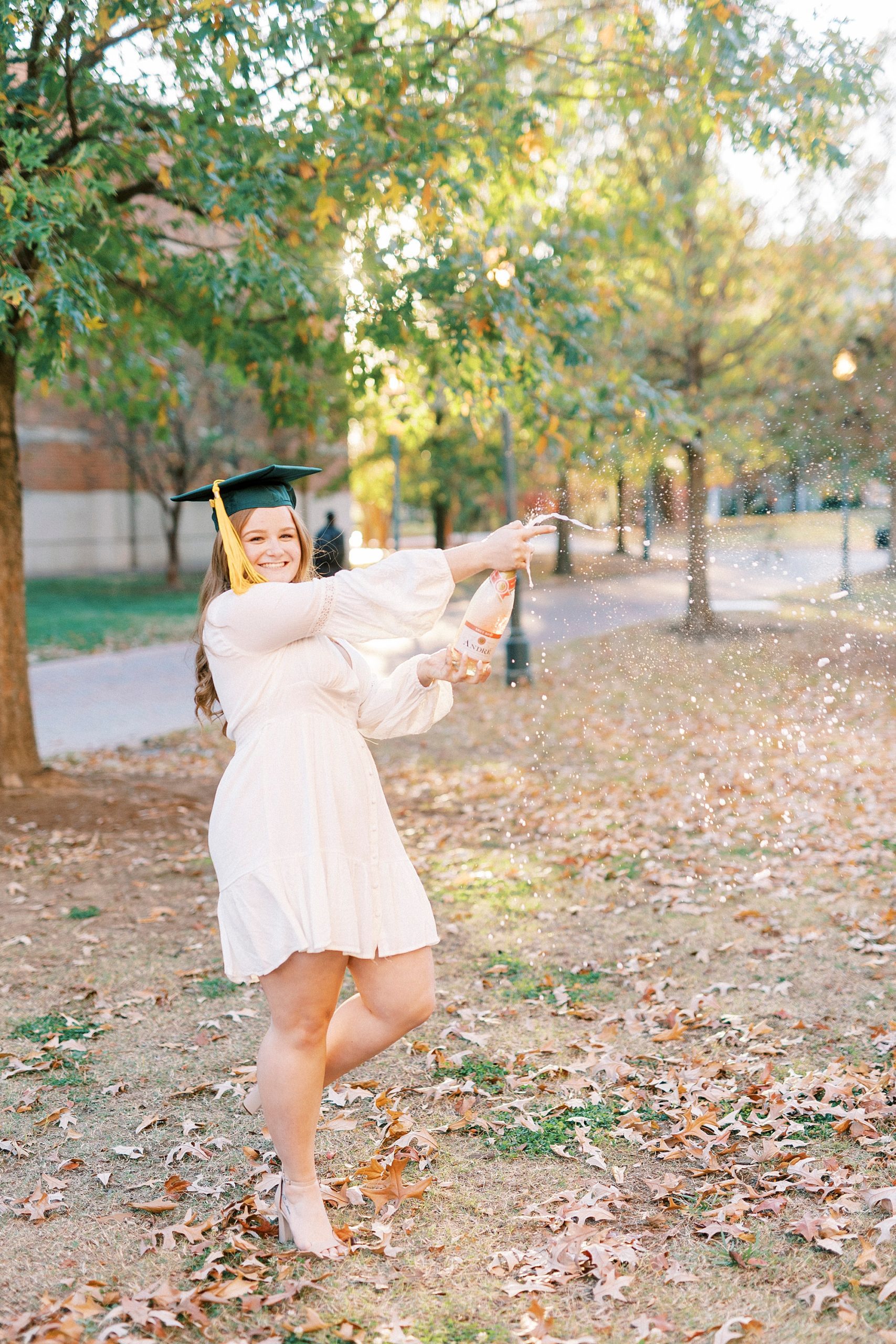 graduate in white dress with green cap pops champagne on lawn at UNC Charlotte