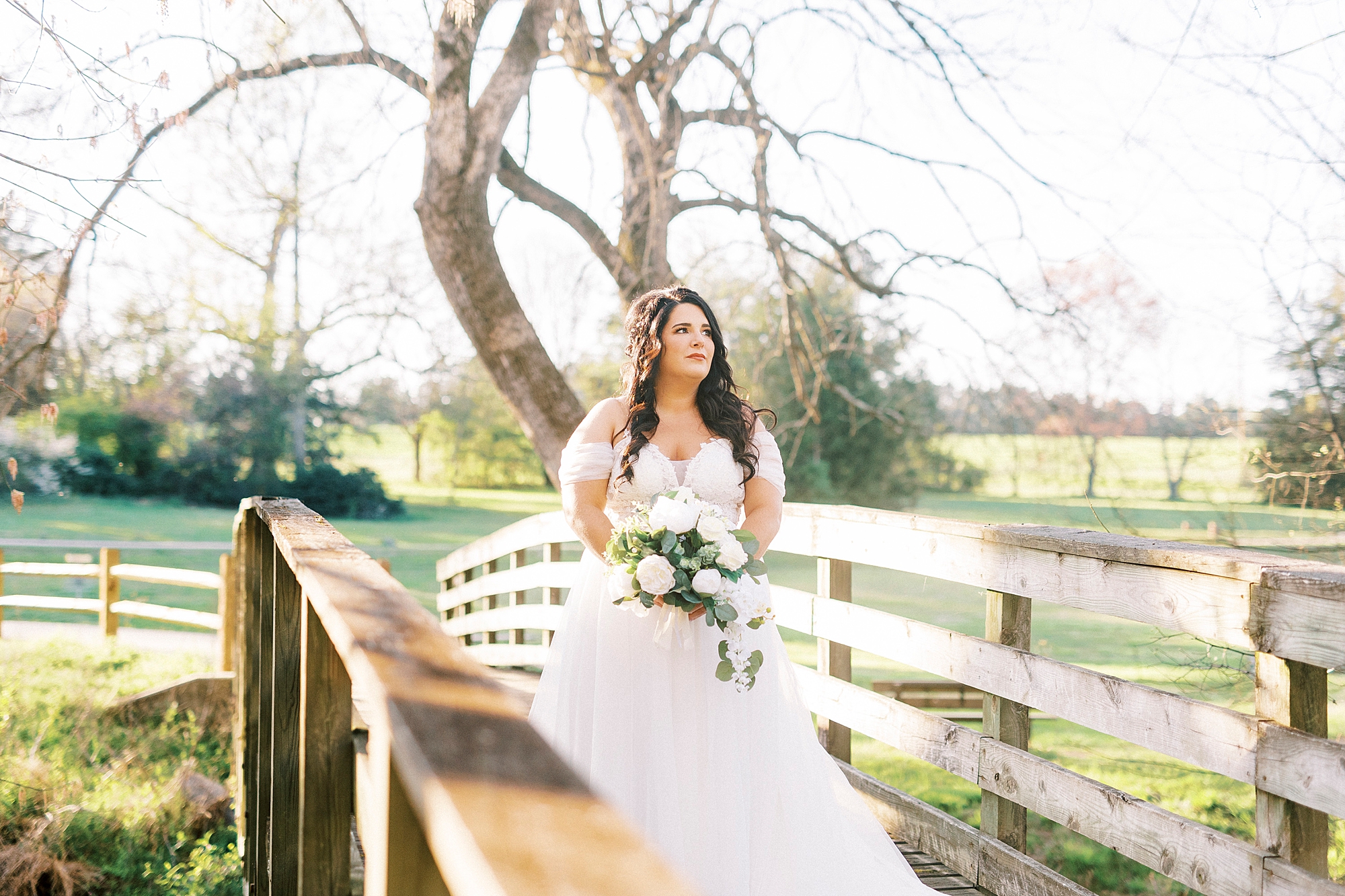 bride with long brown hair holds all-white bouquet on wooden bridge