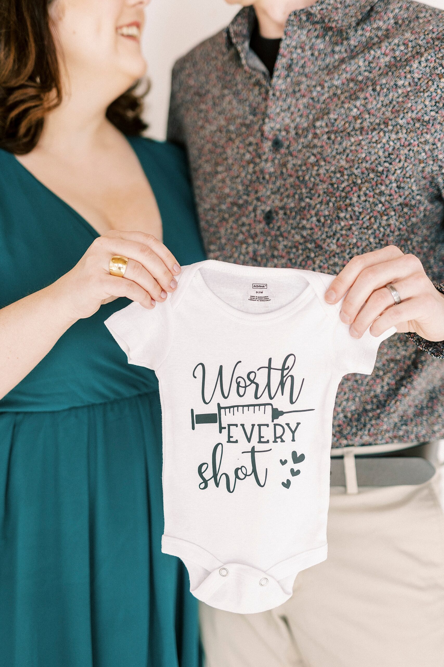 parents hold baby onesie with IVF needles saying "worth every shot"