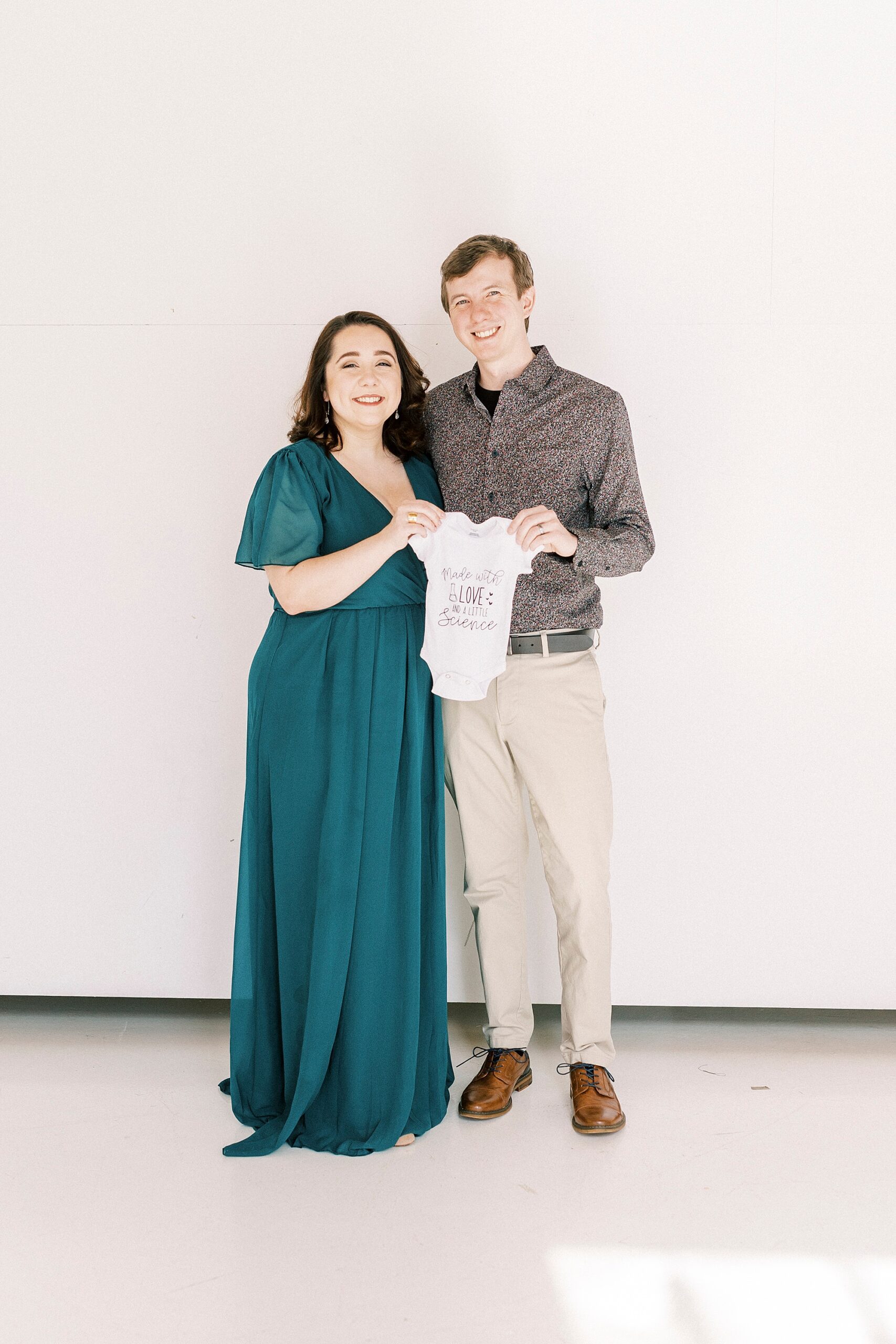 husband and wife hug together during studio pregnancy announcement in Concord NC holding baby onesie