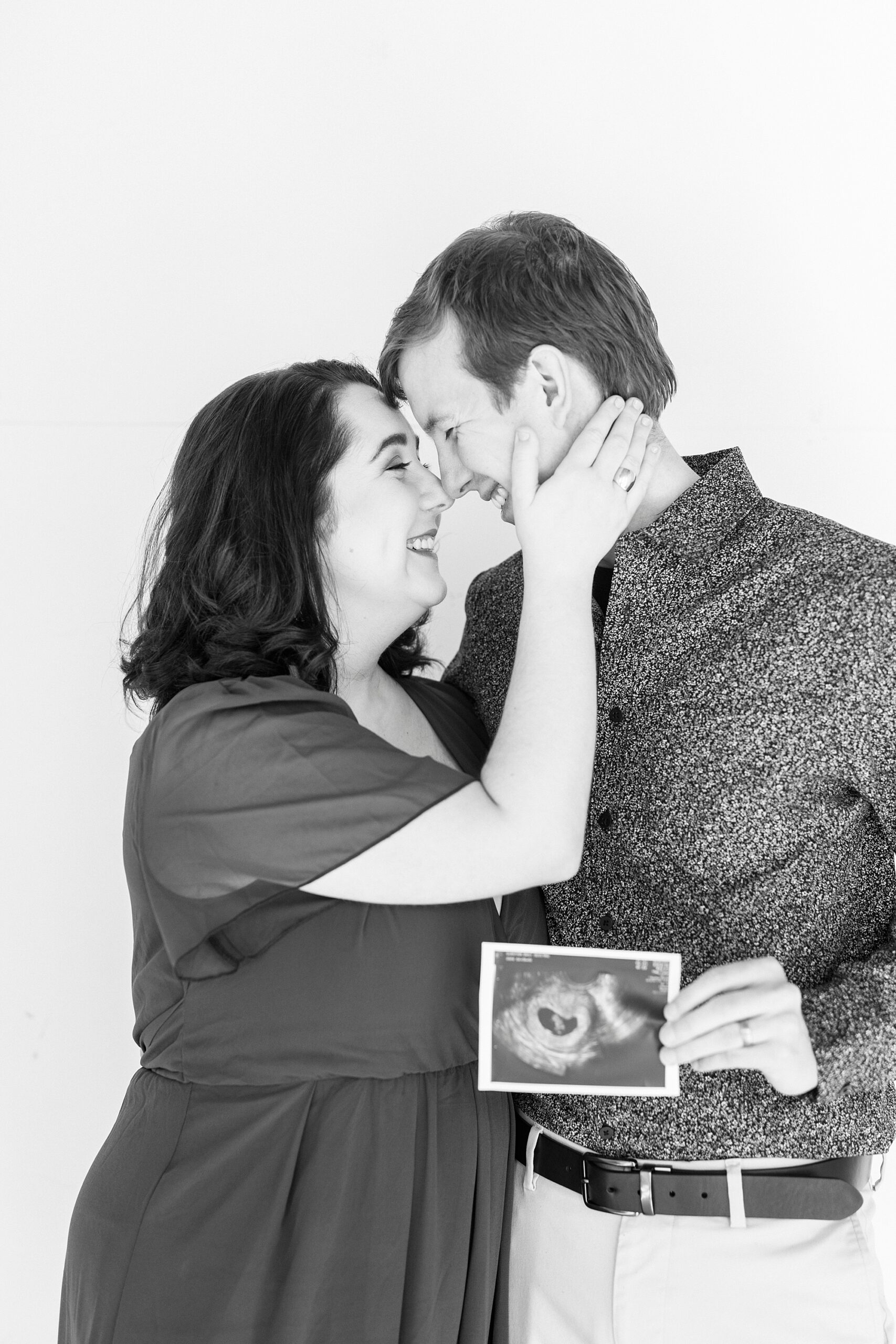 married couple leans heads together holding sonogram during studio pregnancy announcement in Concord NC