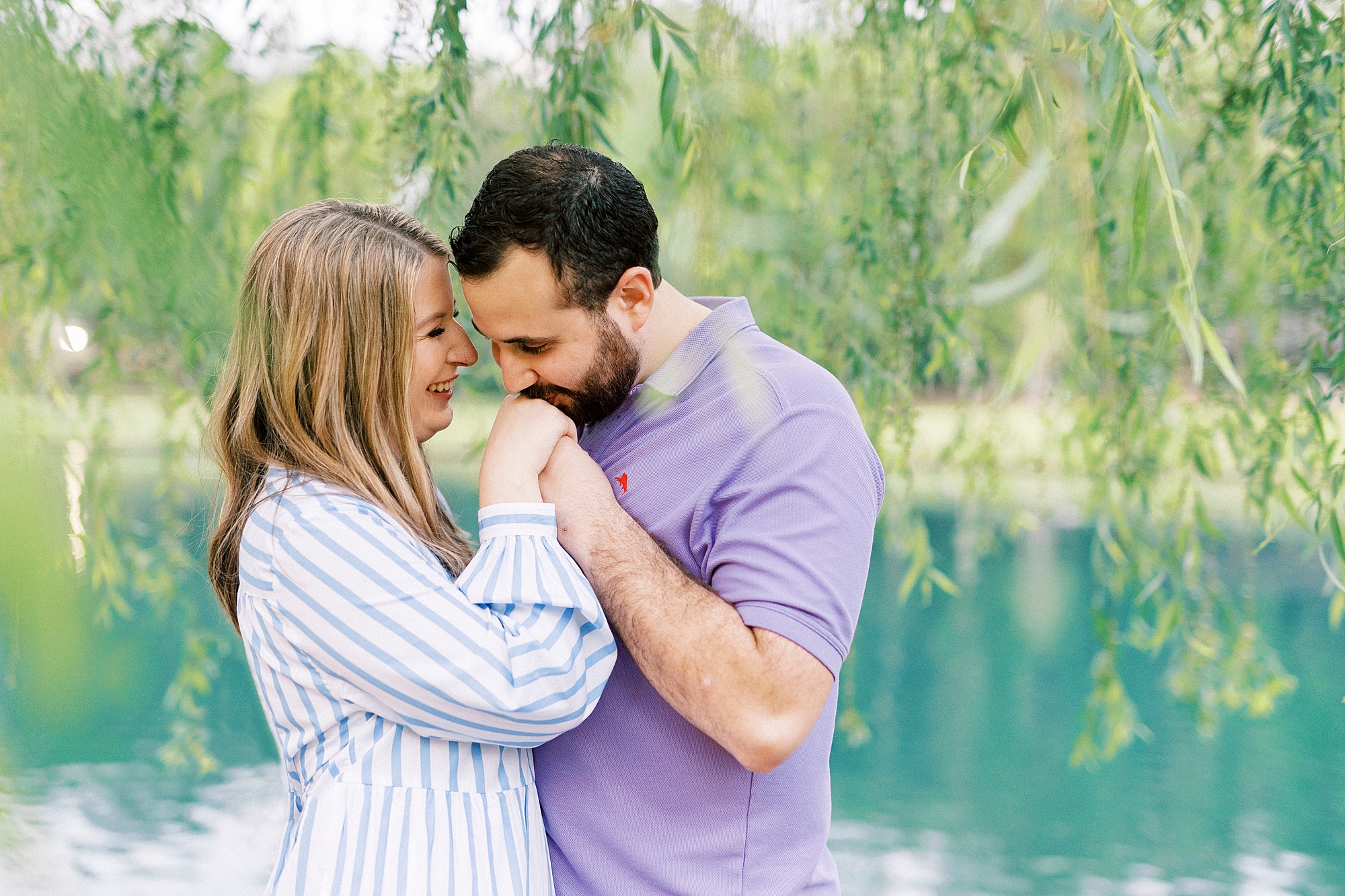 groom in purple polo kisses bride's hand under weeping willow at Freedom Park