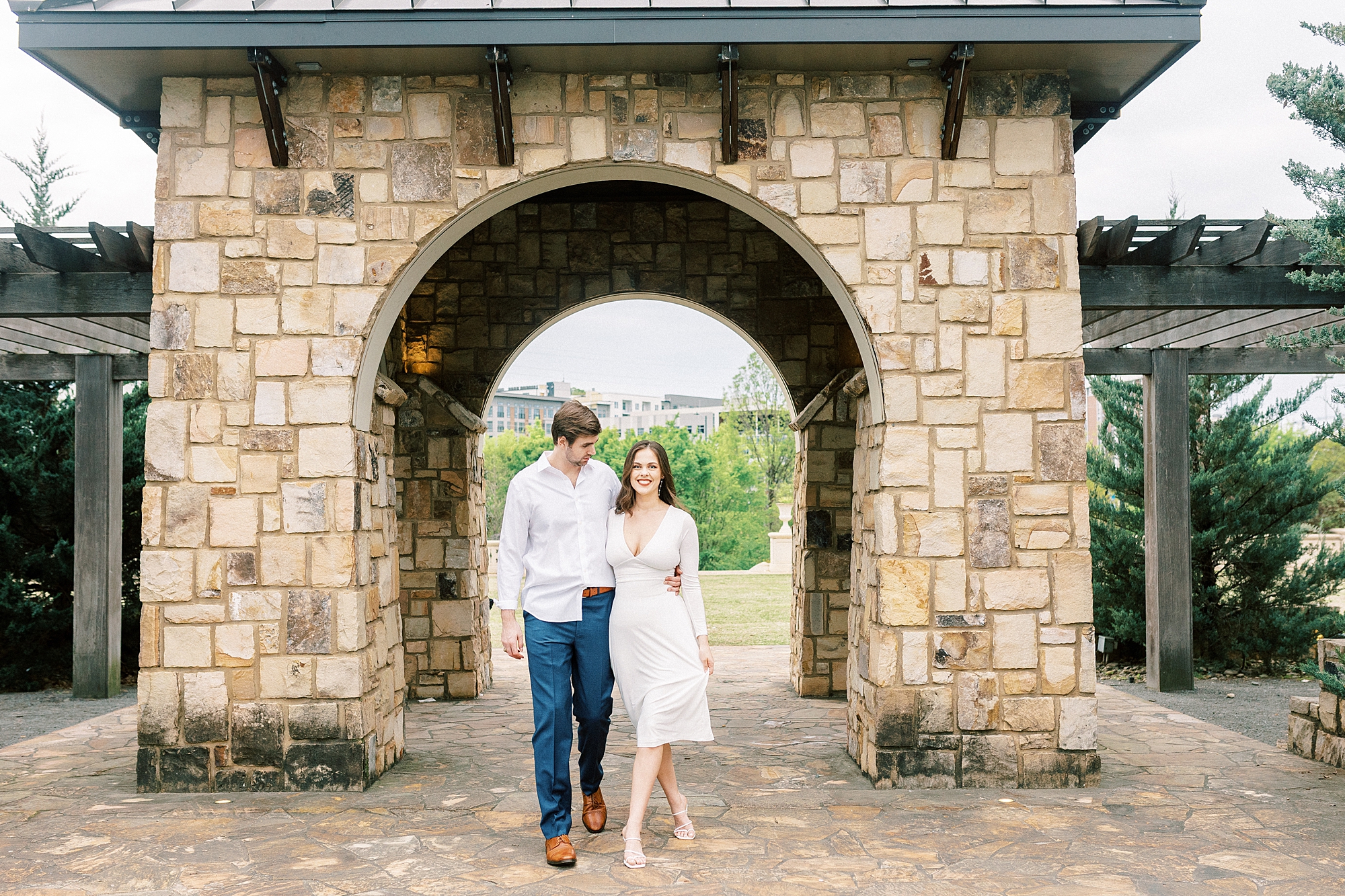 engaged couple walks under stone arbor at Midtown Park