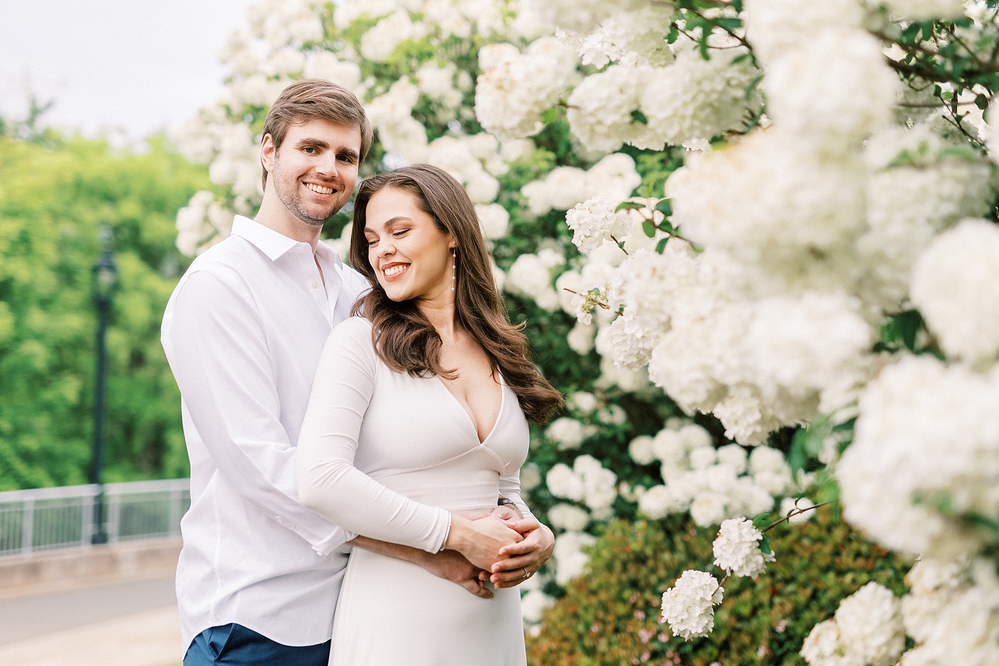 man hugs woman in white dress from behind with white flowers at Midtown Park