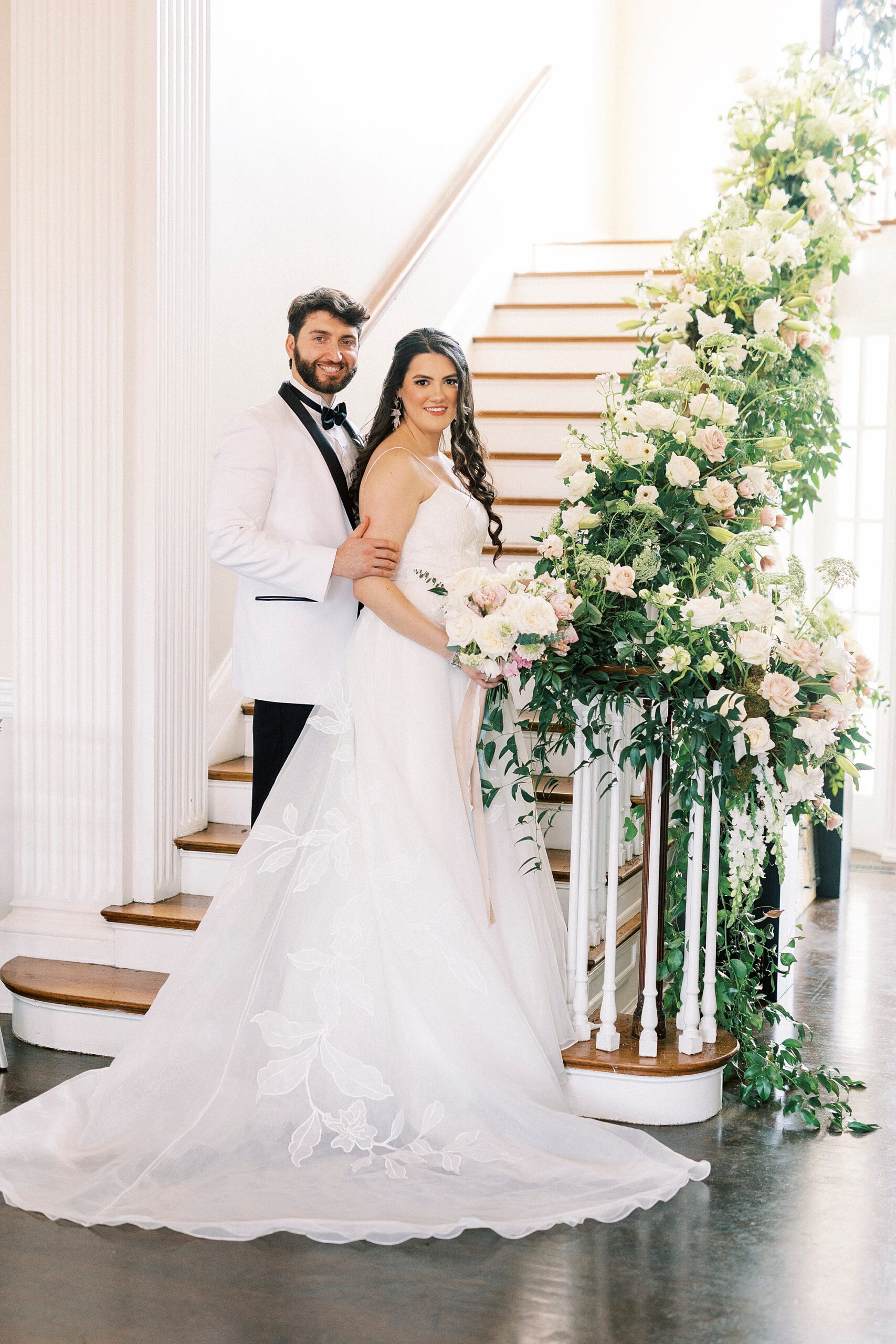 groom in white tux jacket holds bride's hips standing at base of staircase in Separk Mansion