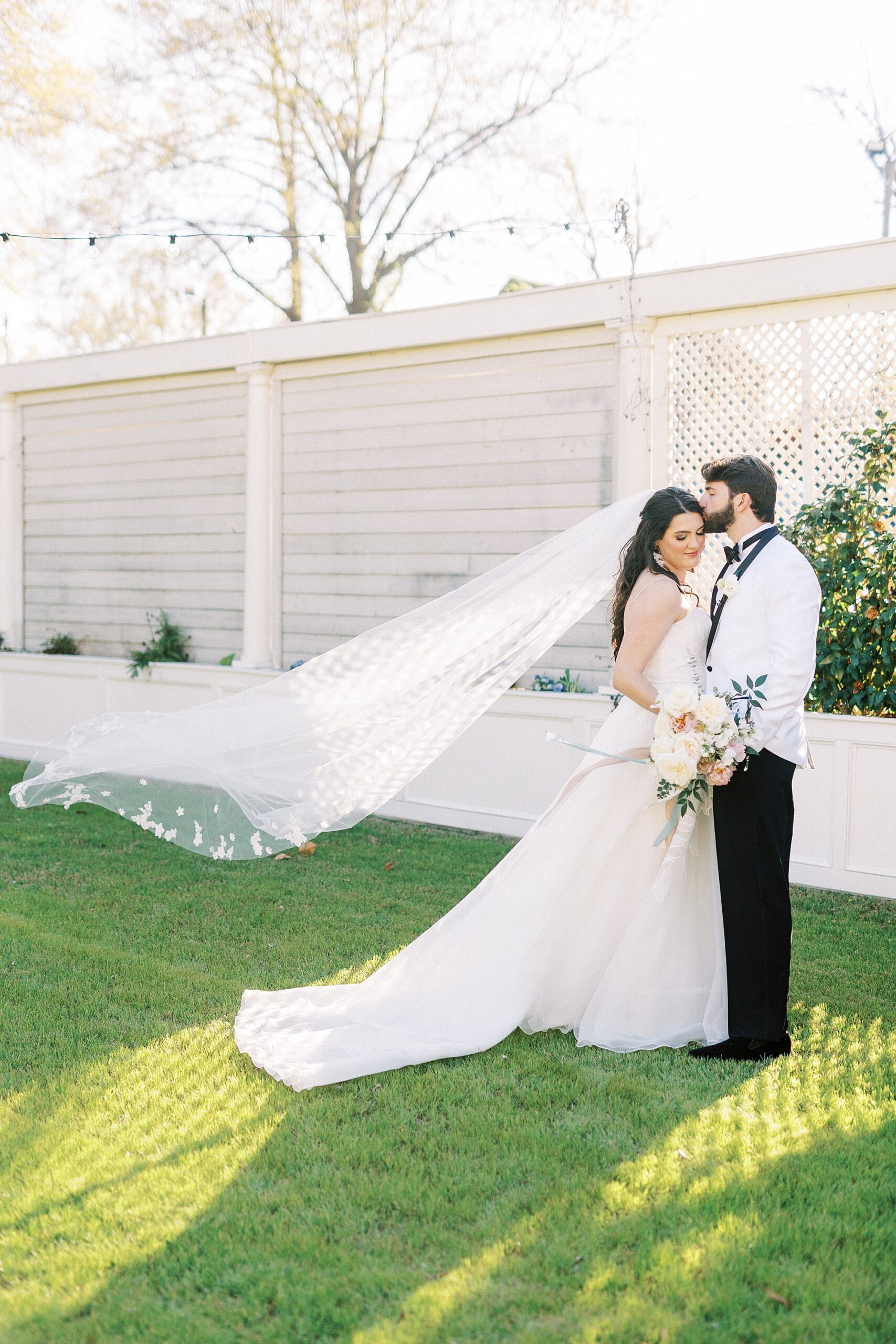 groom kisses bride's forehead with veil floating behind them 