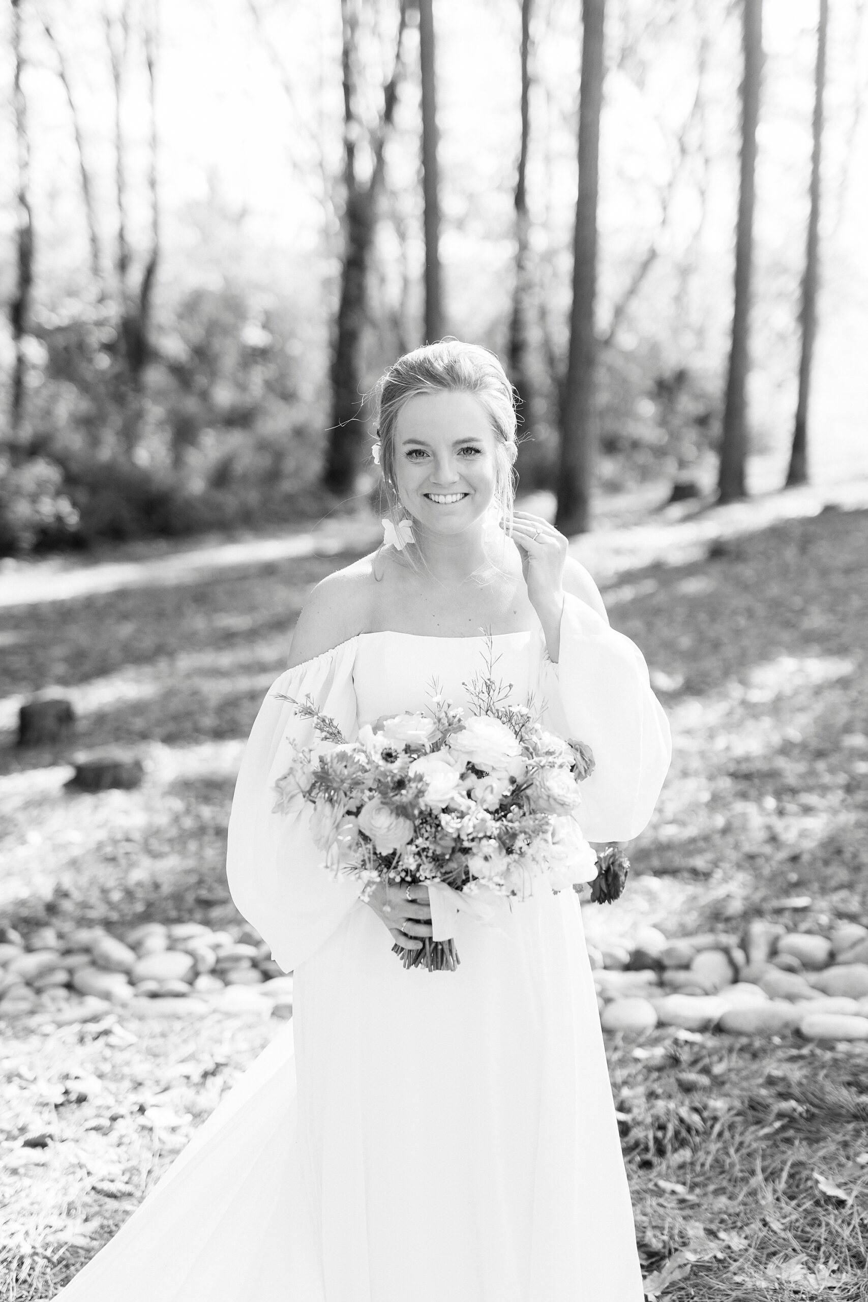 bride in off-the-shoulder boho inspired wedding gown holds bouquet in woods