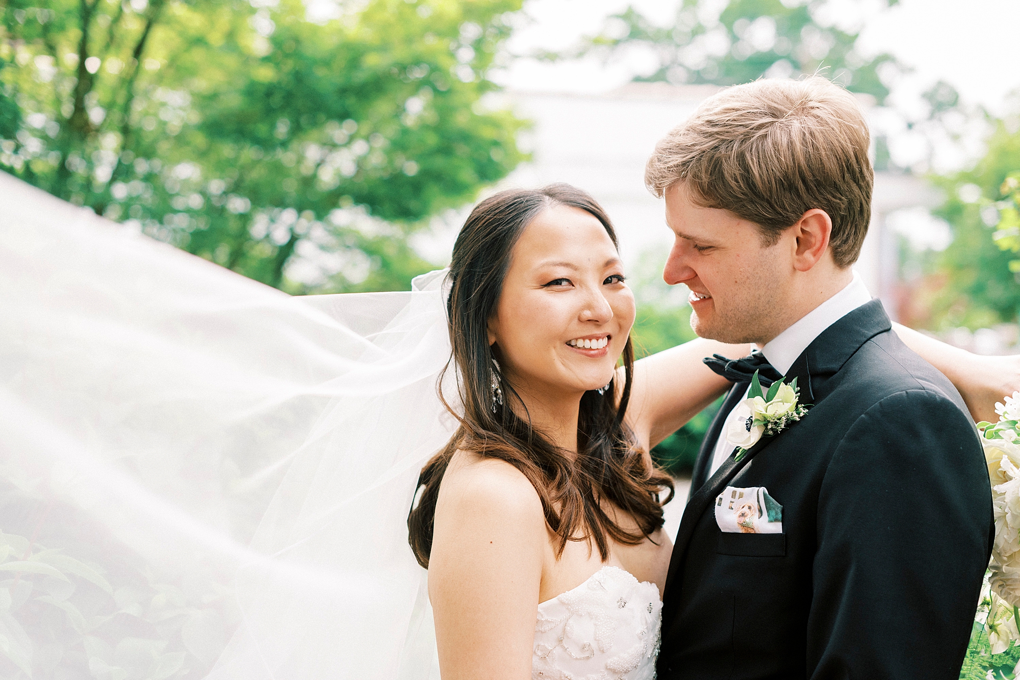 groom looks at bride while she smiles at camera with veil floating behind her